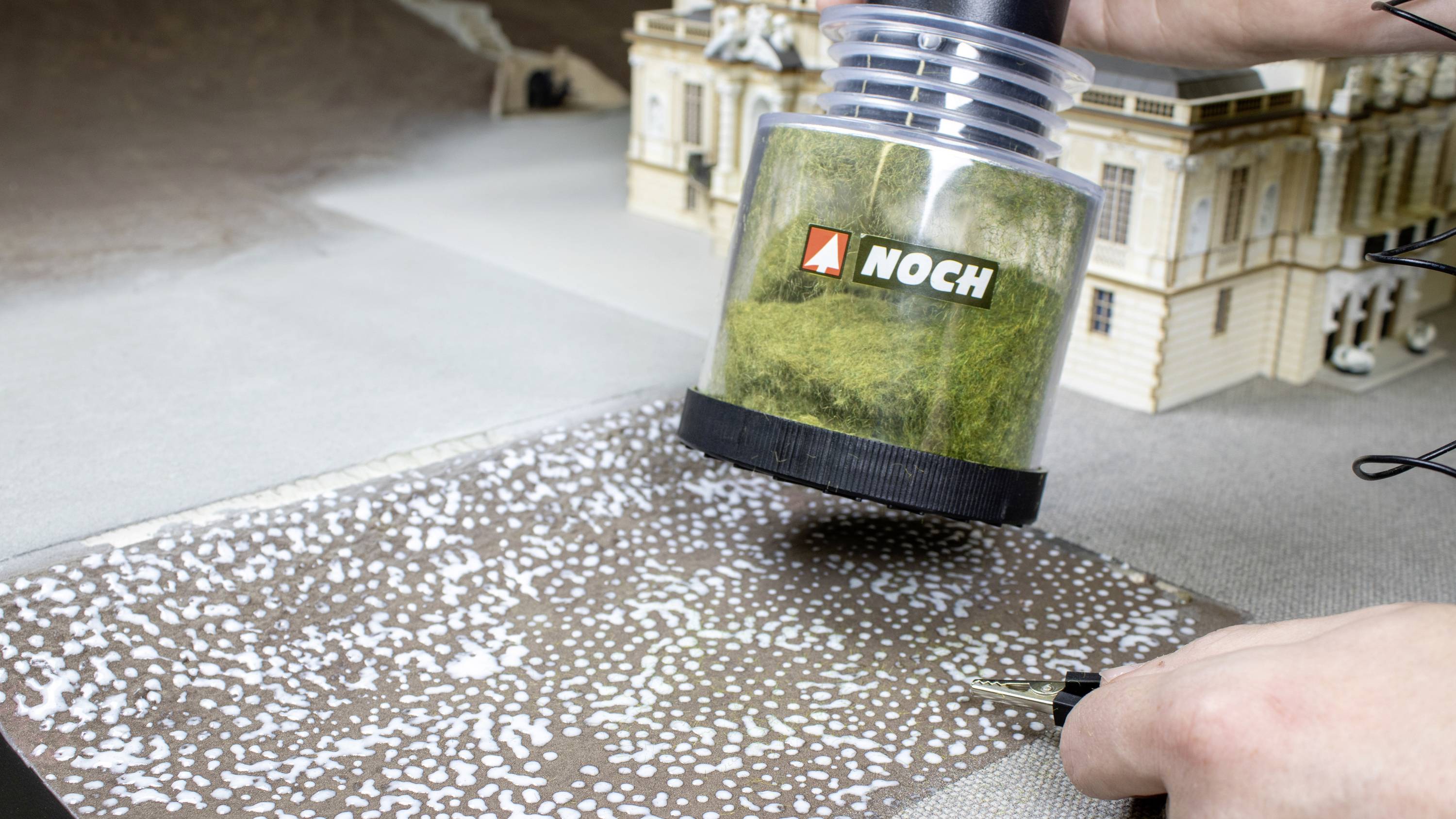 Een persoon gebruikt een gereedschap om groen materiaal aan te brengen op een model-diorama dat een gebouw met een park voorstelt.