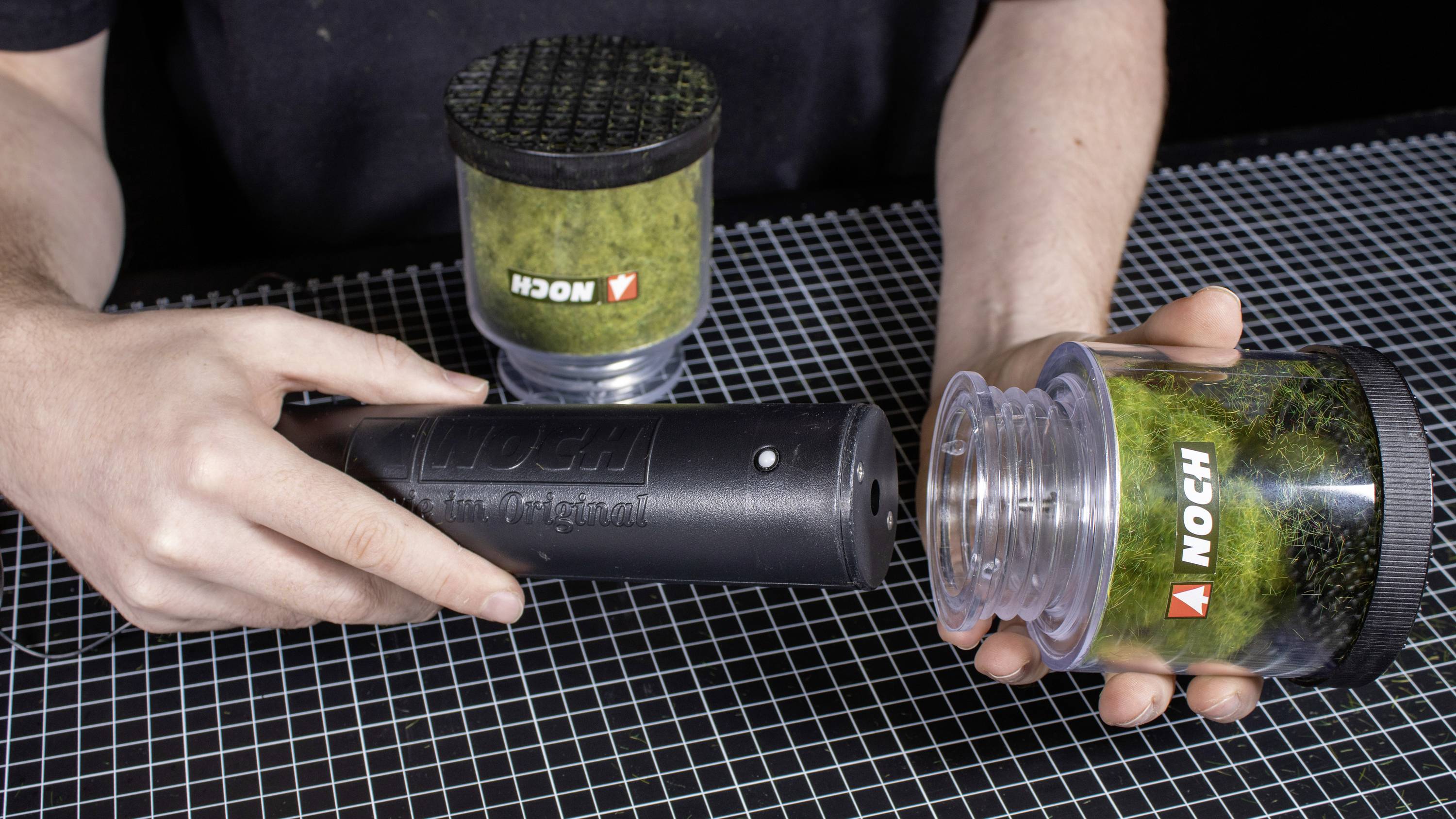 Een persoon demonstreert hoe men mosvezel uit een container aanbrengt met een elektrostatische applicator om modellandschappen te creëren.