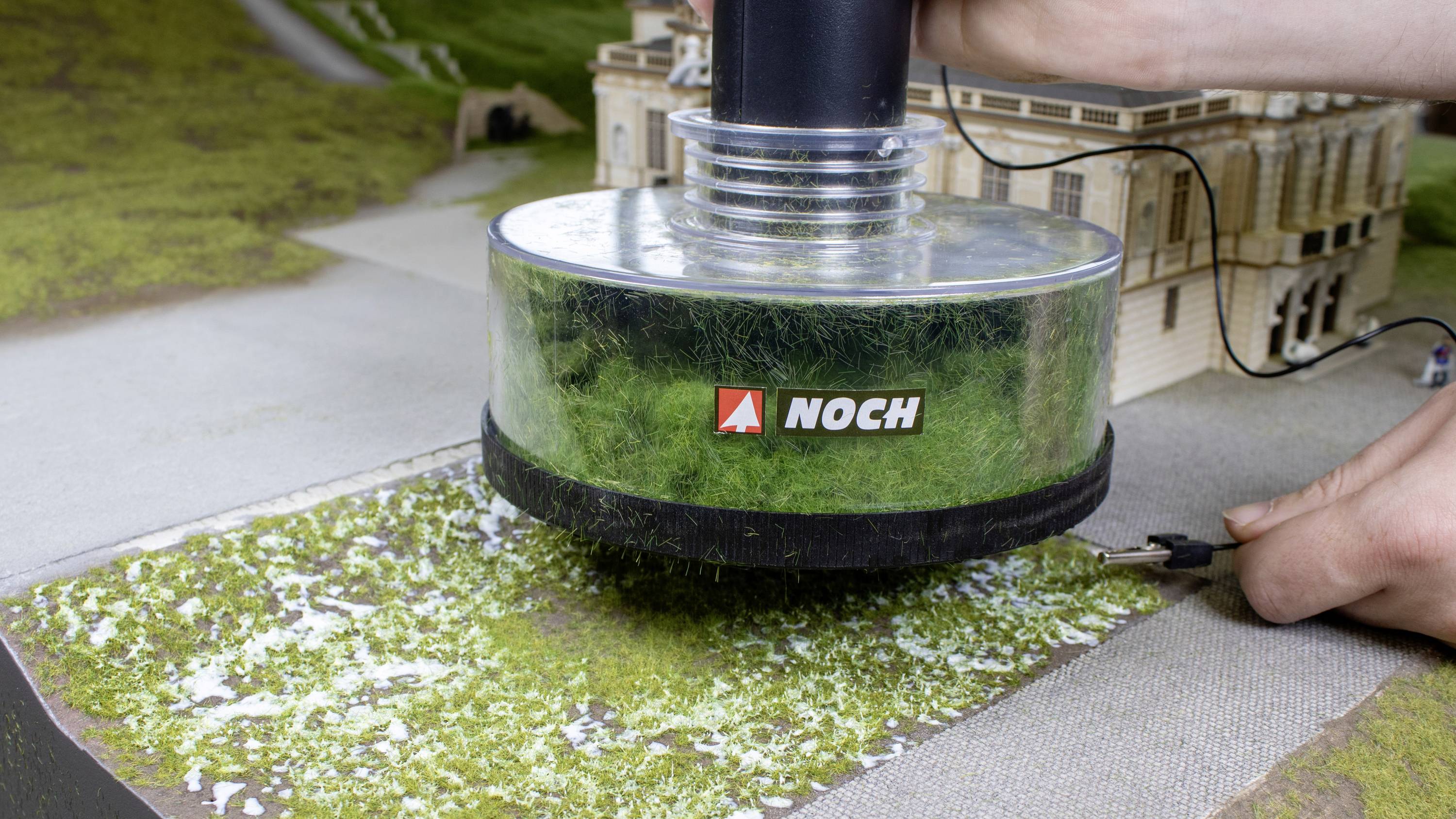 Een hand houdt een apparaat vast met het opschrift 'NOCH', dat gras aanbrengt op een model van een gebouwencomplex.
