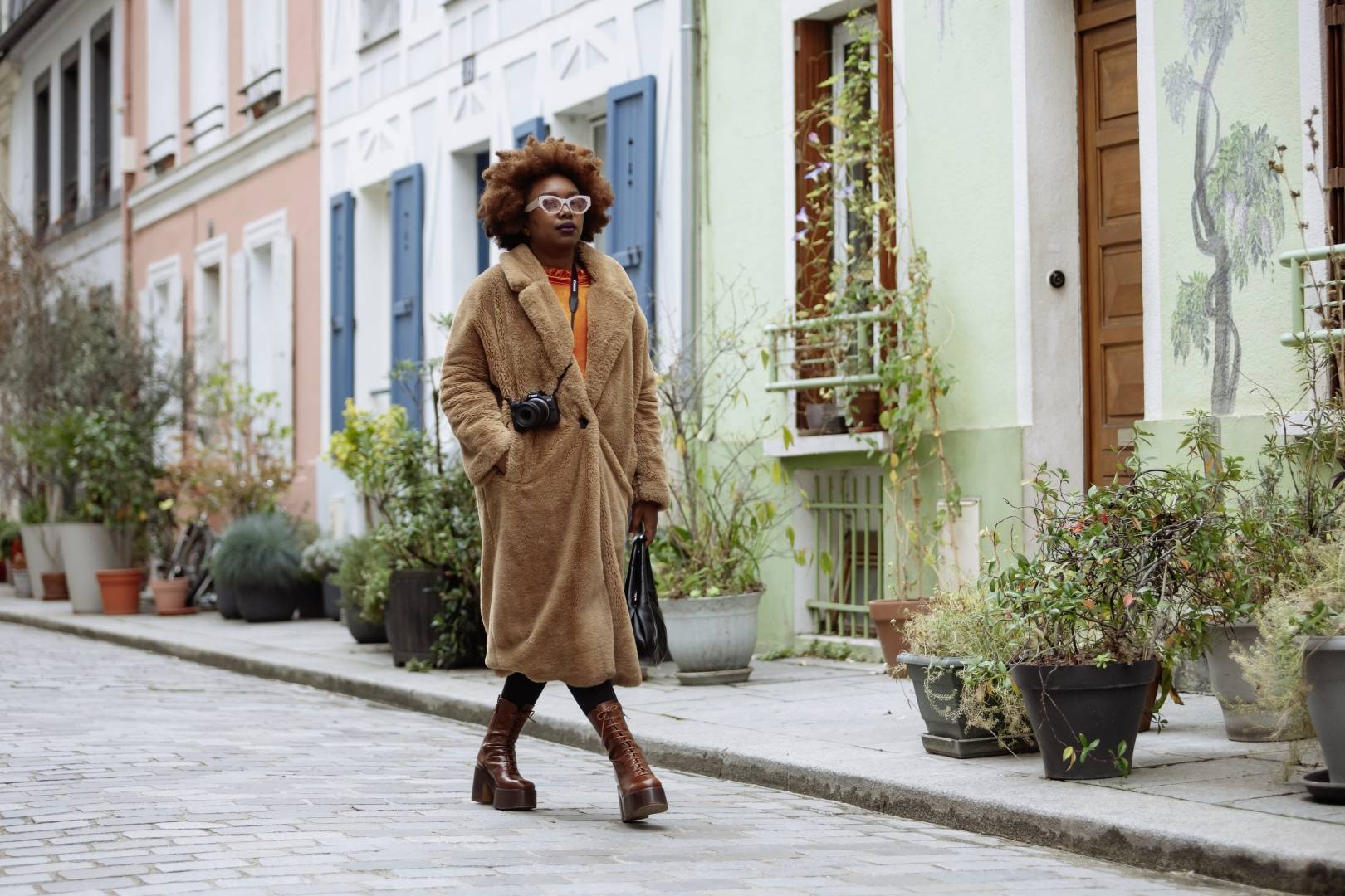 Een persoon met een bruine jas en een camera om de nek wandelt langs een kleurrijke straatsteeg, omgeven door planten.