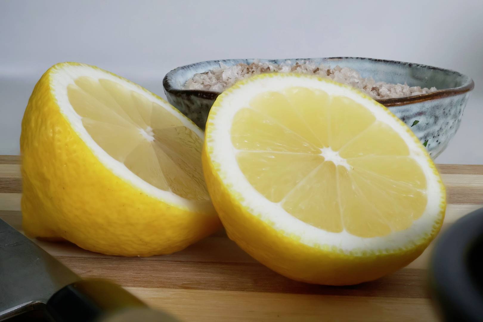 Twee gehalveerde citroenen liggen op een houten plank, met op de achtergrond een schaaltje met grof zout.