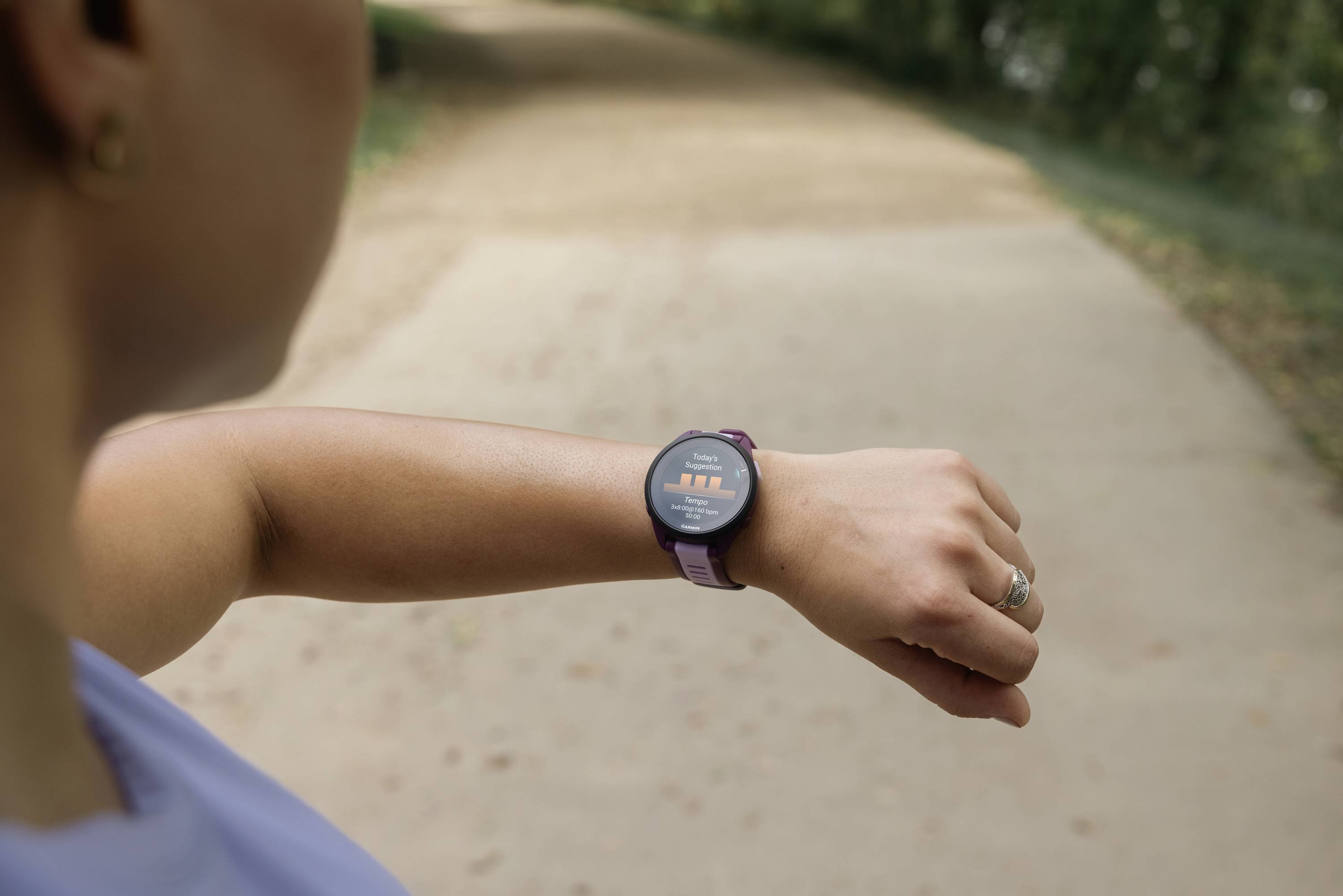 Een persoon kijkt naar een smartwatch terwijl hij/zij buiten op een pad aan het hardlopen is. De horloge toont fitnessgegevens.