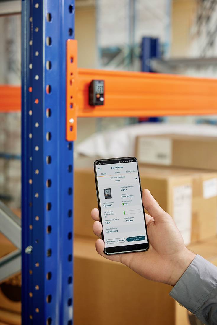 Een persoon houdt een smartphone vast waarop een voorraadbeheersapp wordt weergegeven. Op de achtergrond staan gestapelde dozen in het magazijn.