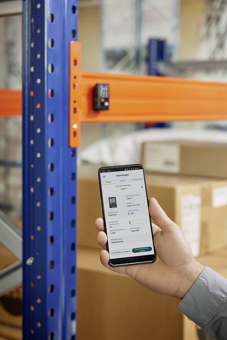 Een persoon houdt een smartphone voor stellingen in een magazijn. Op het scherm wordt een voorraadbeheersapp weergegeven.