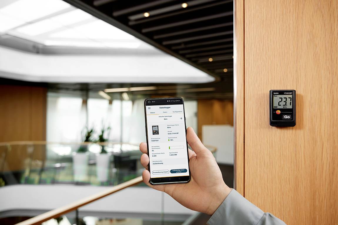 Een persoon houdt een smartphone vast met een app voor ruimtebewaking, naast een digitale thermostaat die is ingesteld op 23,7 graden Celsius.