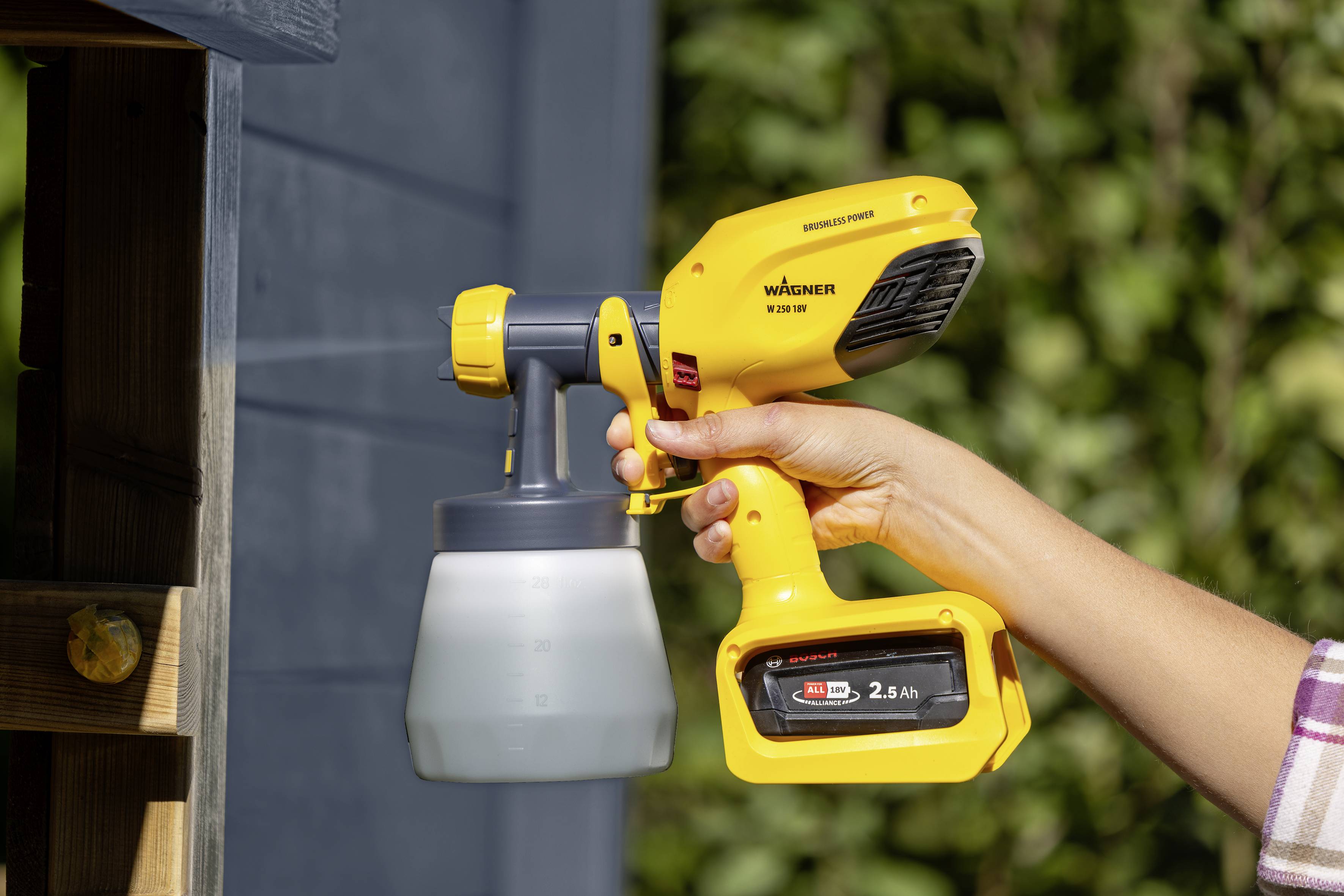 'Een persoon houdt een geel, draadloos verfspuitapparaat vast en spuit daarmee een blauwe houten oppervlakte buiten, omringd door planten.'