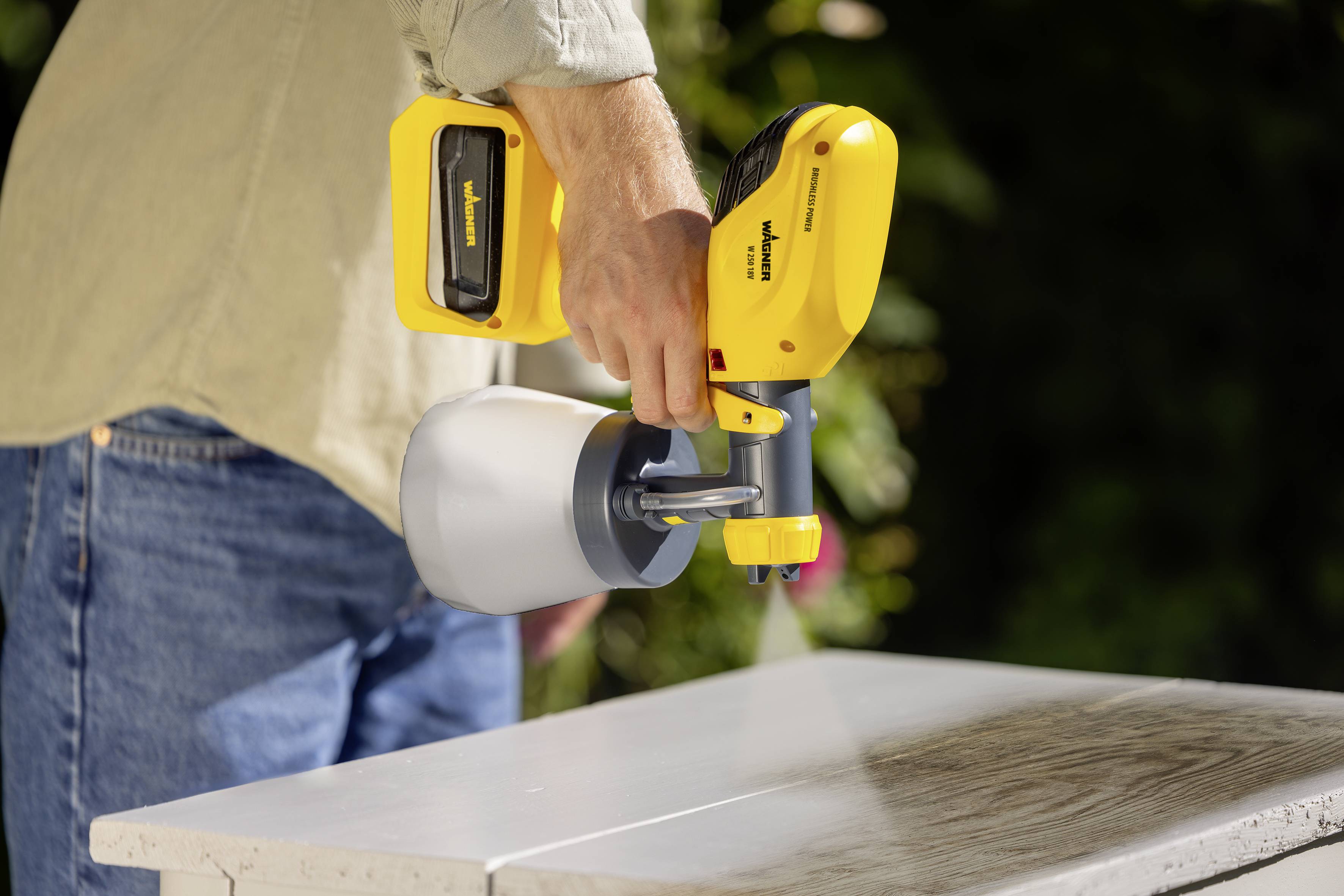 Een persoon houdt een gele verfspuitpistool en schildert een houten oppervlak. De focus ligt op de toepassing van de verf.