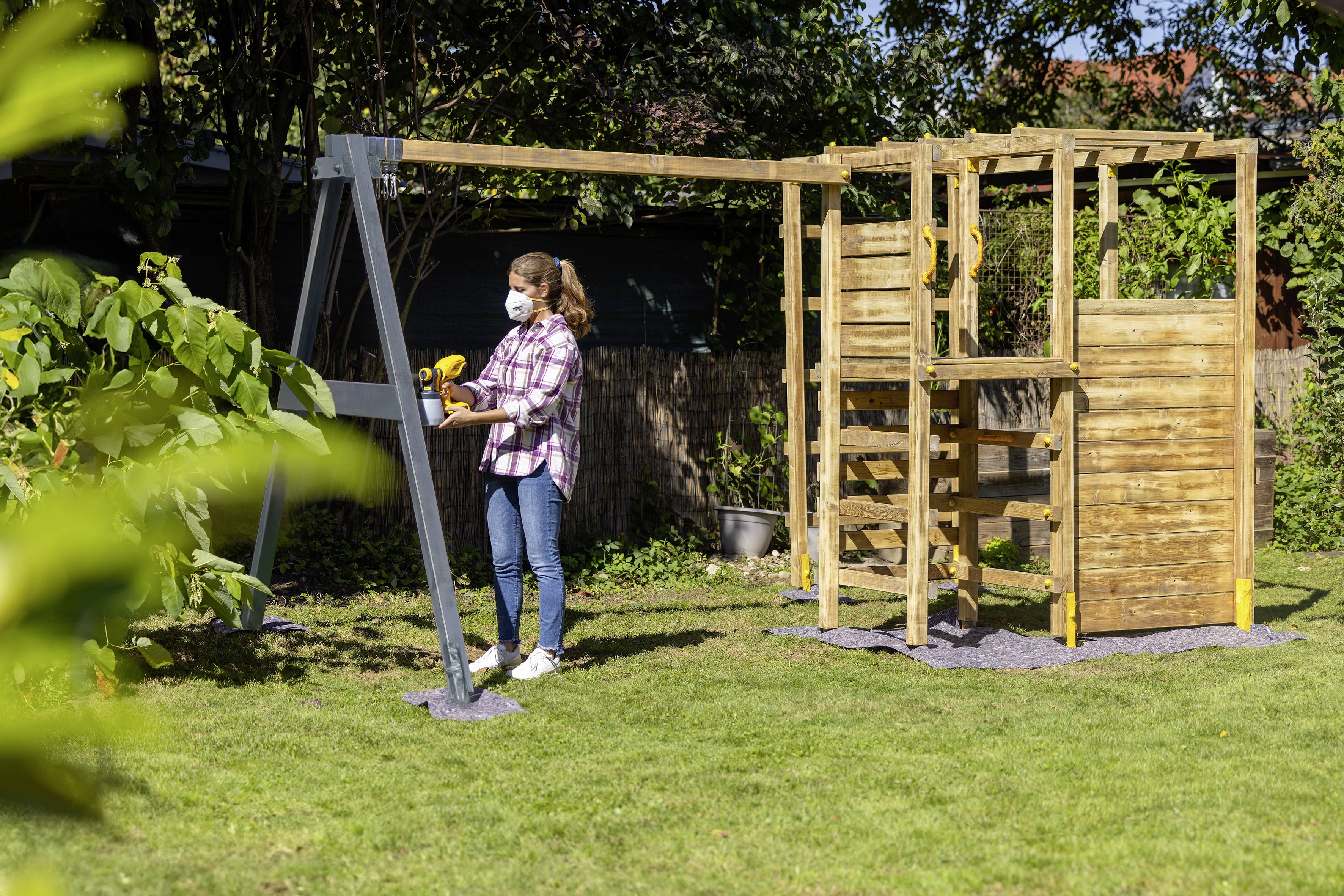Een vrouw staat in de tuin en monteert een houten klimstellage. Ze draagt een beschermingsmasker en gebruikt een boormachine.