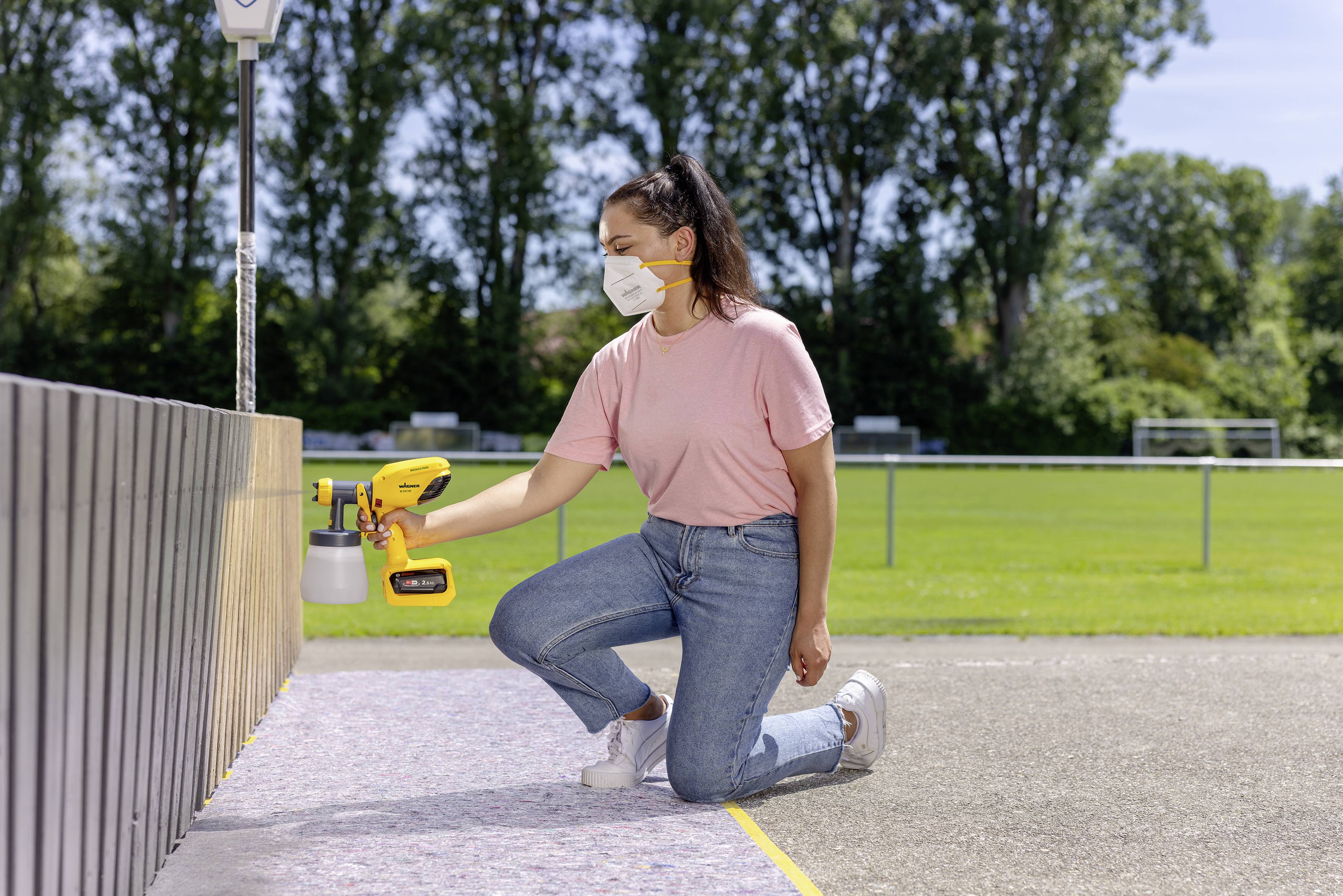 Een persoon met een gezichtsmasker knielt op de grond en gebruikt een geel spuitapparaat om een houten oppervlak buiten te verven.