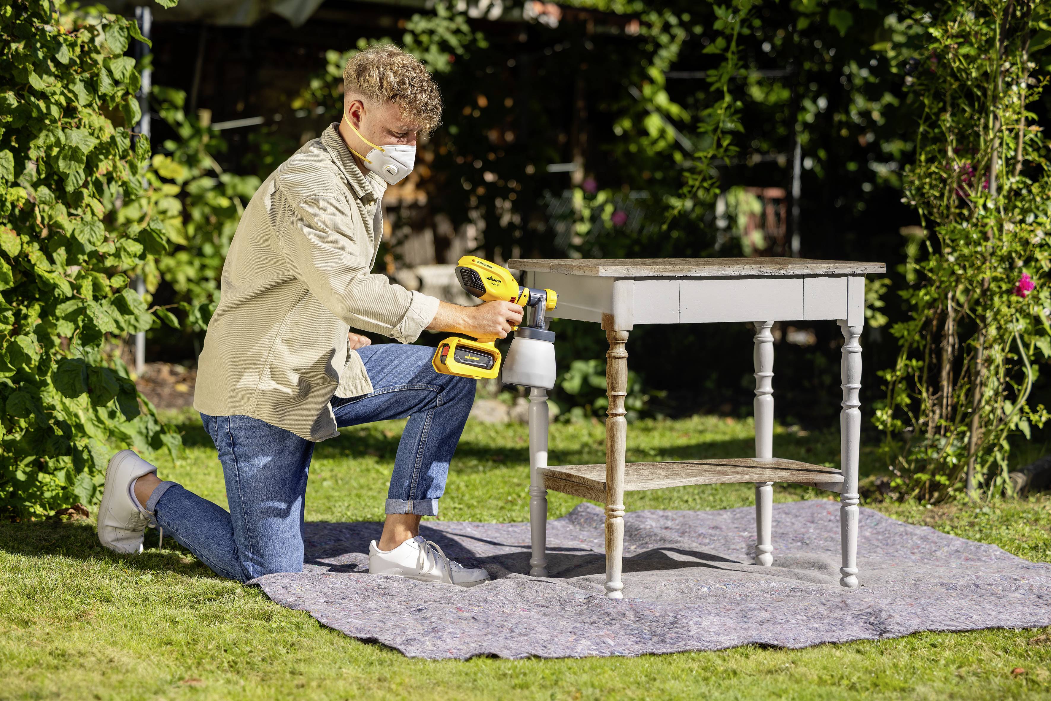 Een persoon buiten schildert een oude tafel met een gele verfspuitpistool. Ze draagt een masker en knielt op een blauw doek op het gras.