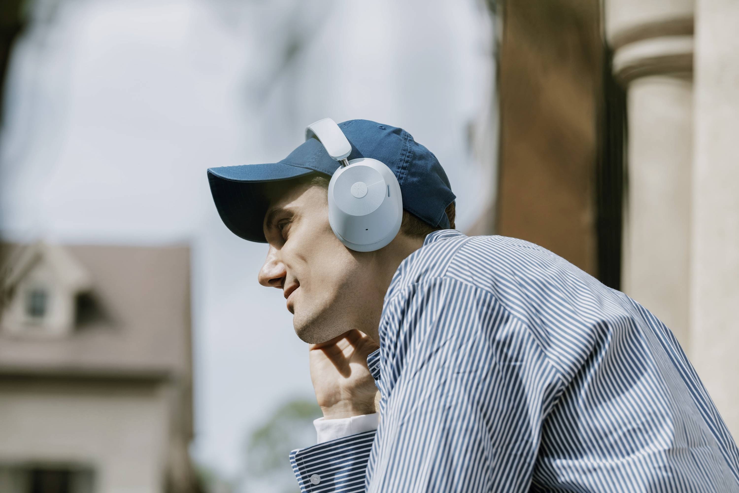Een man in een gestreept shirt en blauwe pet luistert muziek met koptelefoons en leunt ontspannen tegen een muur buiten.