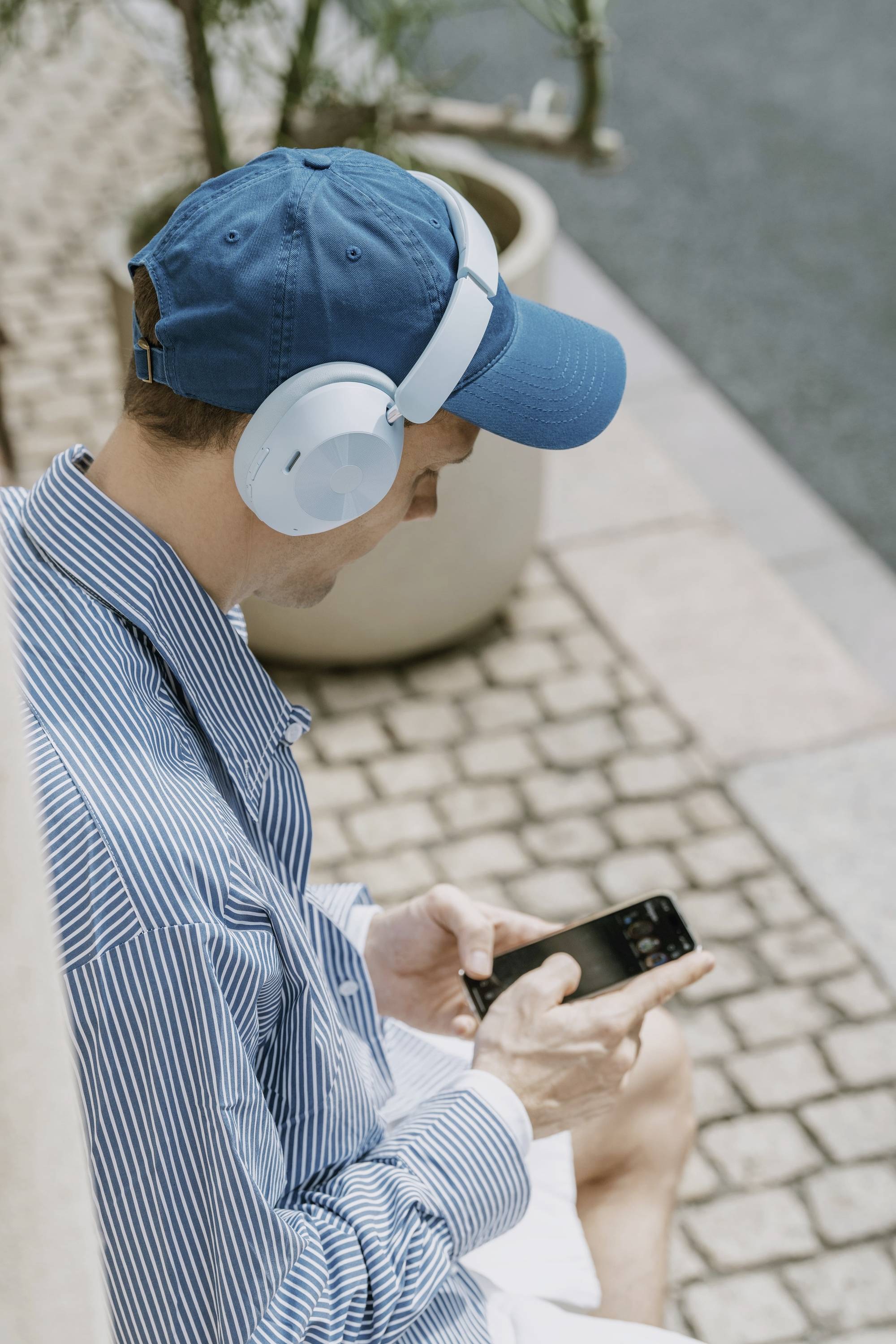 Een man zit buiten, draagt een gestreept shirt, een blauwe pet en een koptelefoon. Hij kijkt naar zijn smartphone.