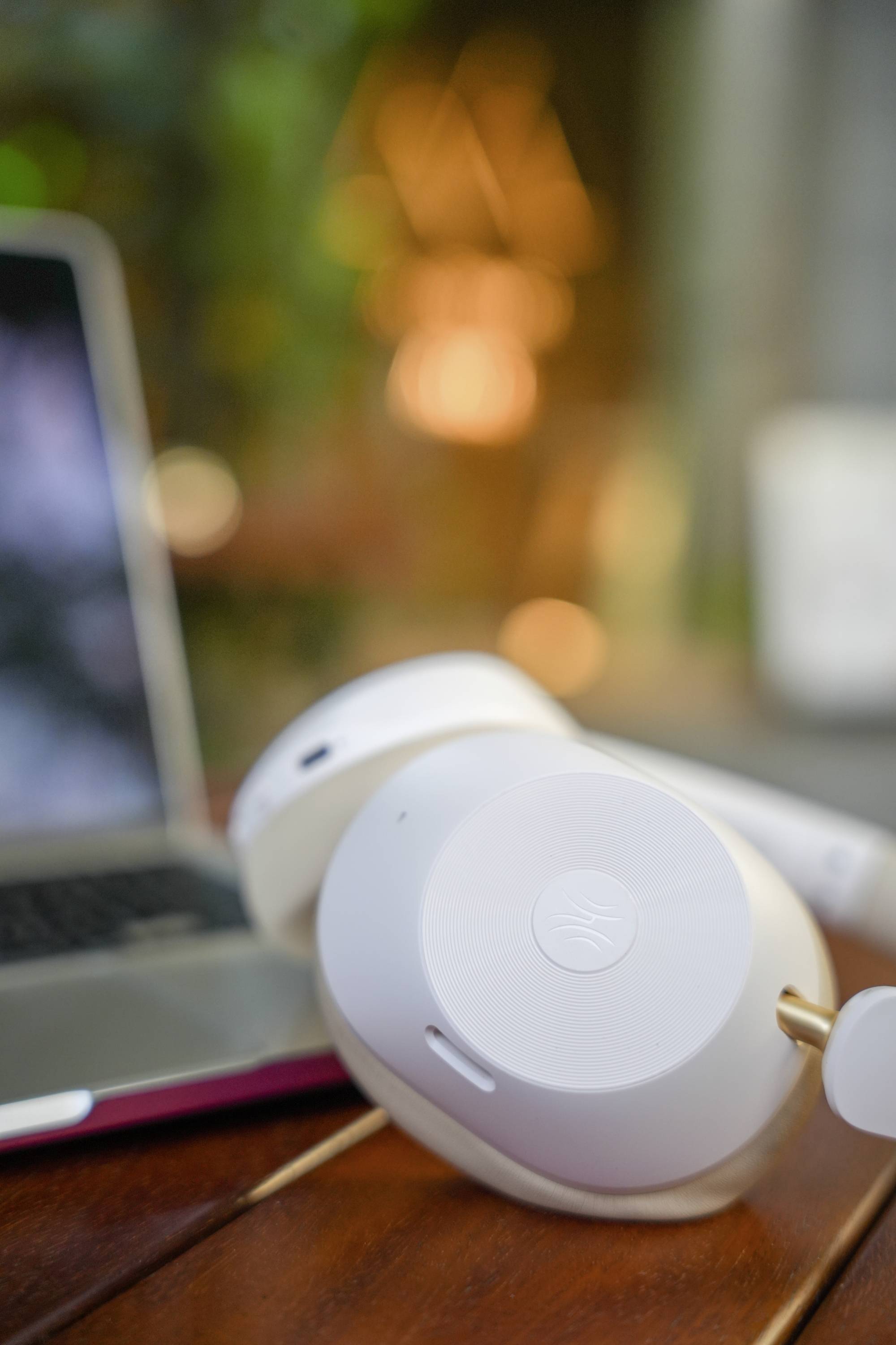 Witte koptelefoon ligt op een houten tafel naast een laptop. De achtergrond is wazig en toont gekleurde lichten.