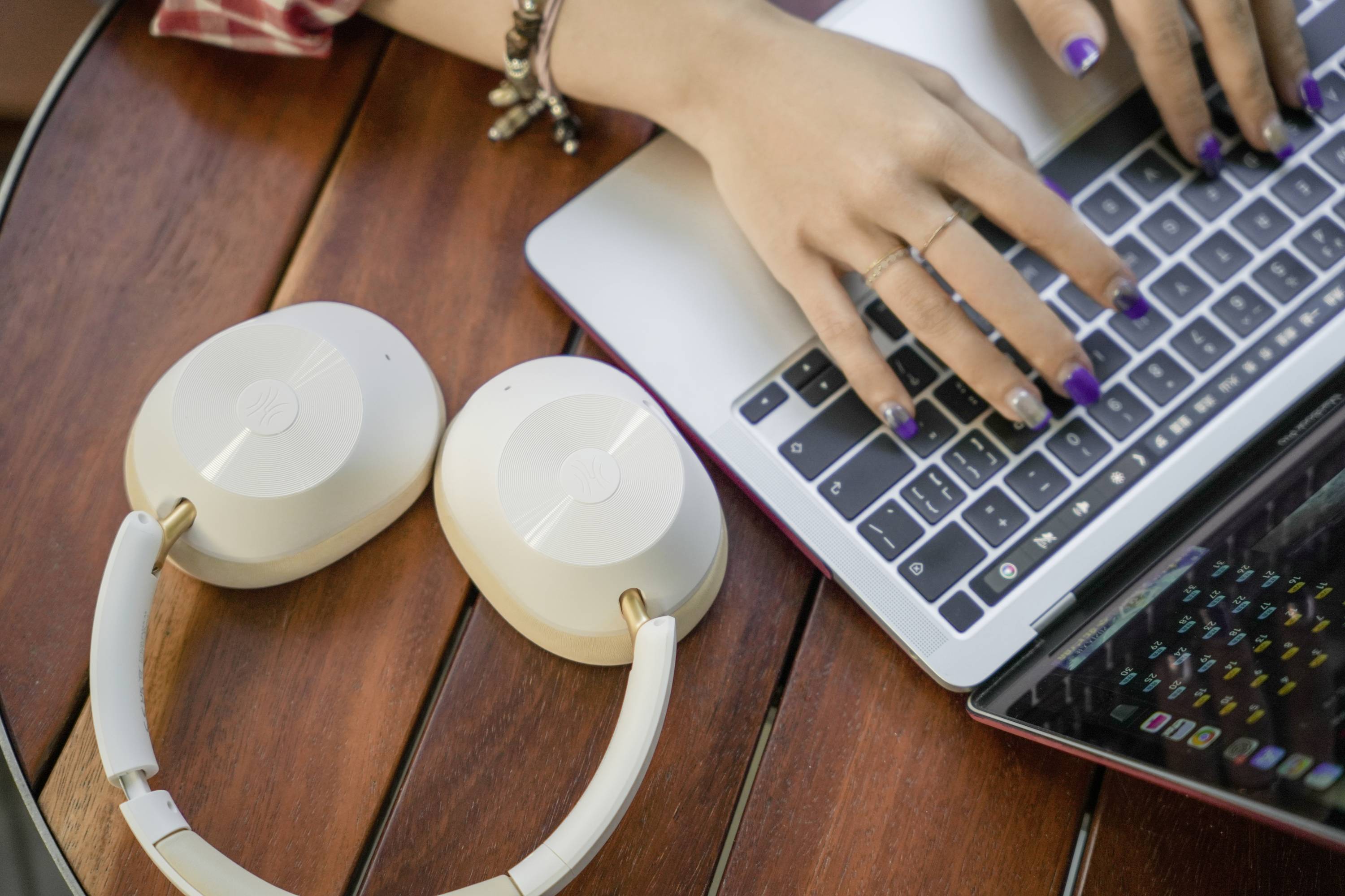 Handen typen op een laptoptoetsenbord, naast een paar witte koptelefoons op een houten tafel.
