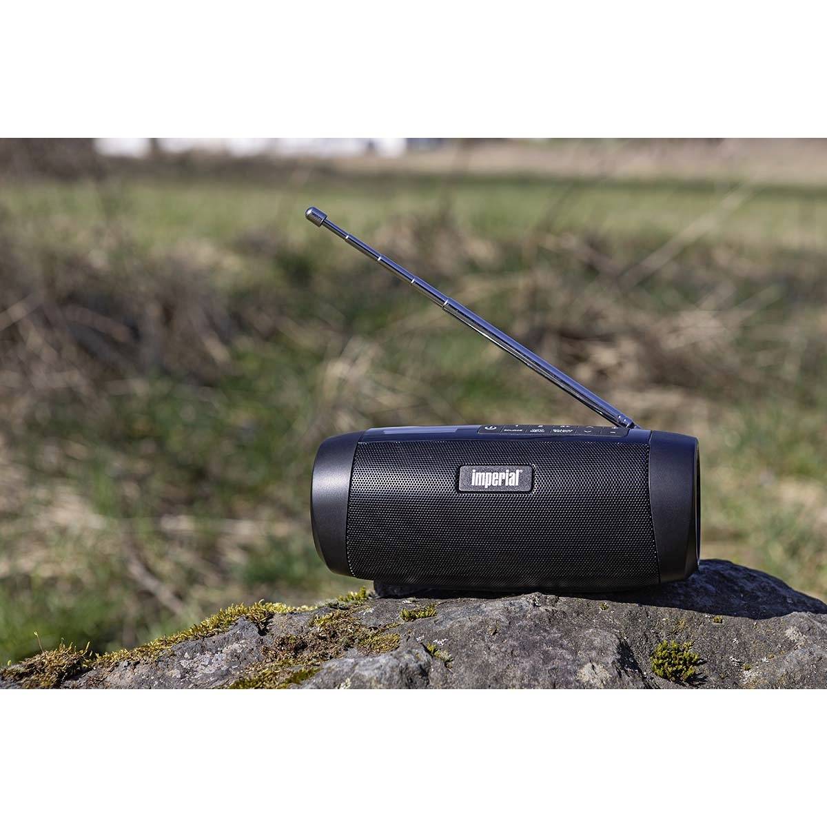 Een draagbare radio met uitgetrokken antenne staat op een rots in de buitenlucht. Op de achtergrond zijn gras en een onscherp landschap zichtbaar.