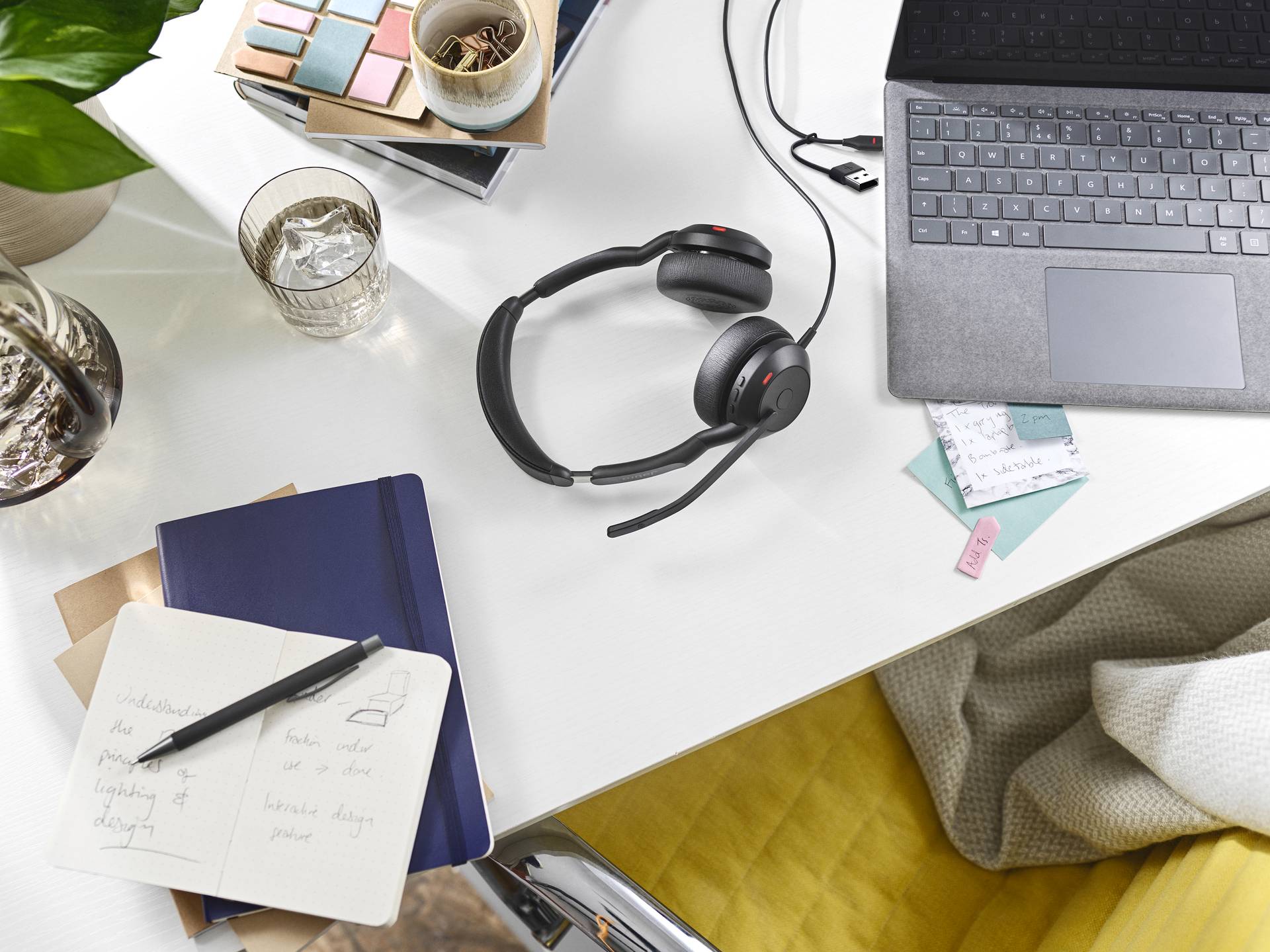 Een bureau met laptop, headset, notitieboek en glas water; een gezellige werkplek met schrijfmaterialen en planten.