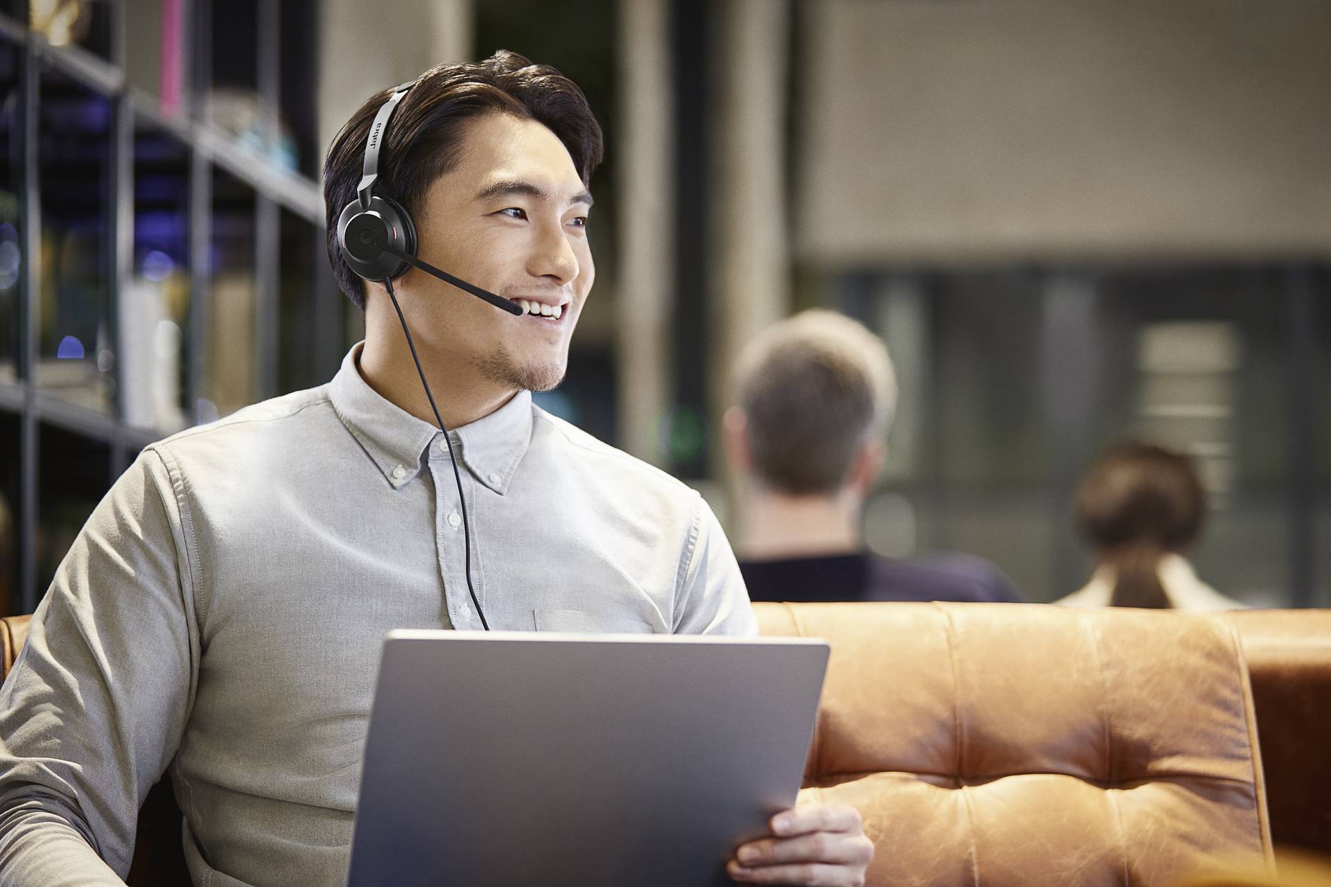 Een man met een headset zit op een bank, houdt een laptop vast en glimlacht terwijl hij werkt op kantoor. Op de achtergrond zijn personen onscherp in beeld.