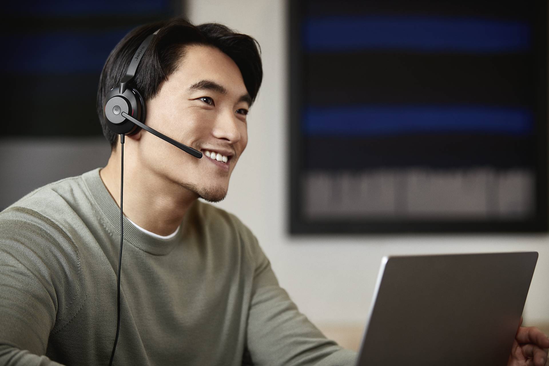 Man met headset en laptop glimlacht tijdens het werk. Achtergrond wazig, toont kantoor- of werkomgeving.
