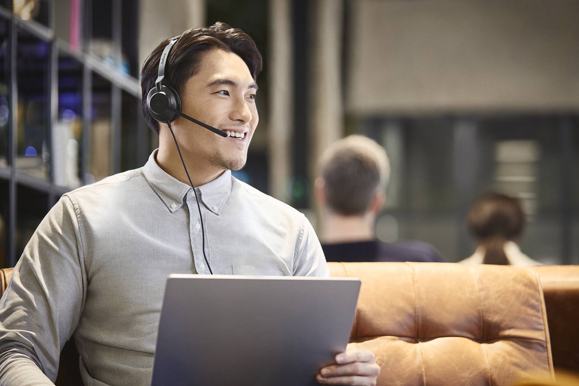 Een man met een headset zit op een lederen bank en houdt een laptop vast. Op de achtergrond zijn wazig personen te zien.