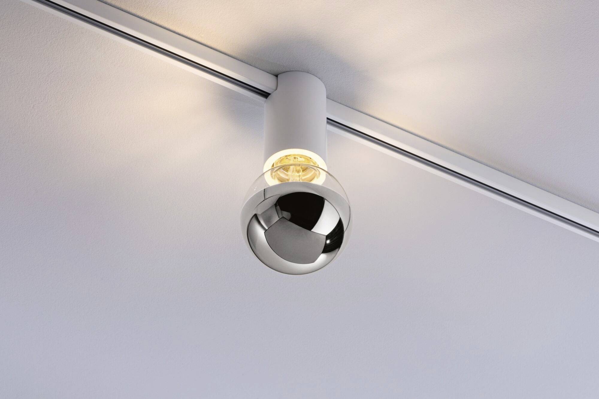 Plafondspot met bol-optica, gemonteerd aan een rail aan het plafond. Straalt warm licht naar beneden, in modern design.