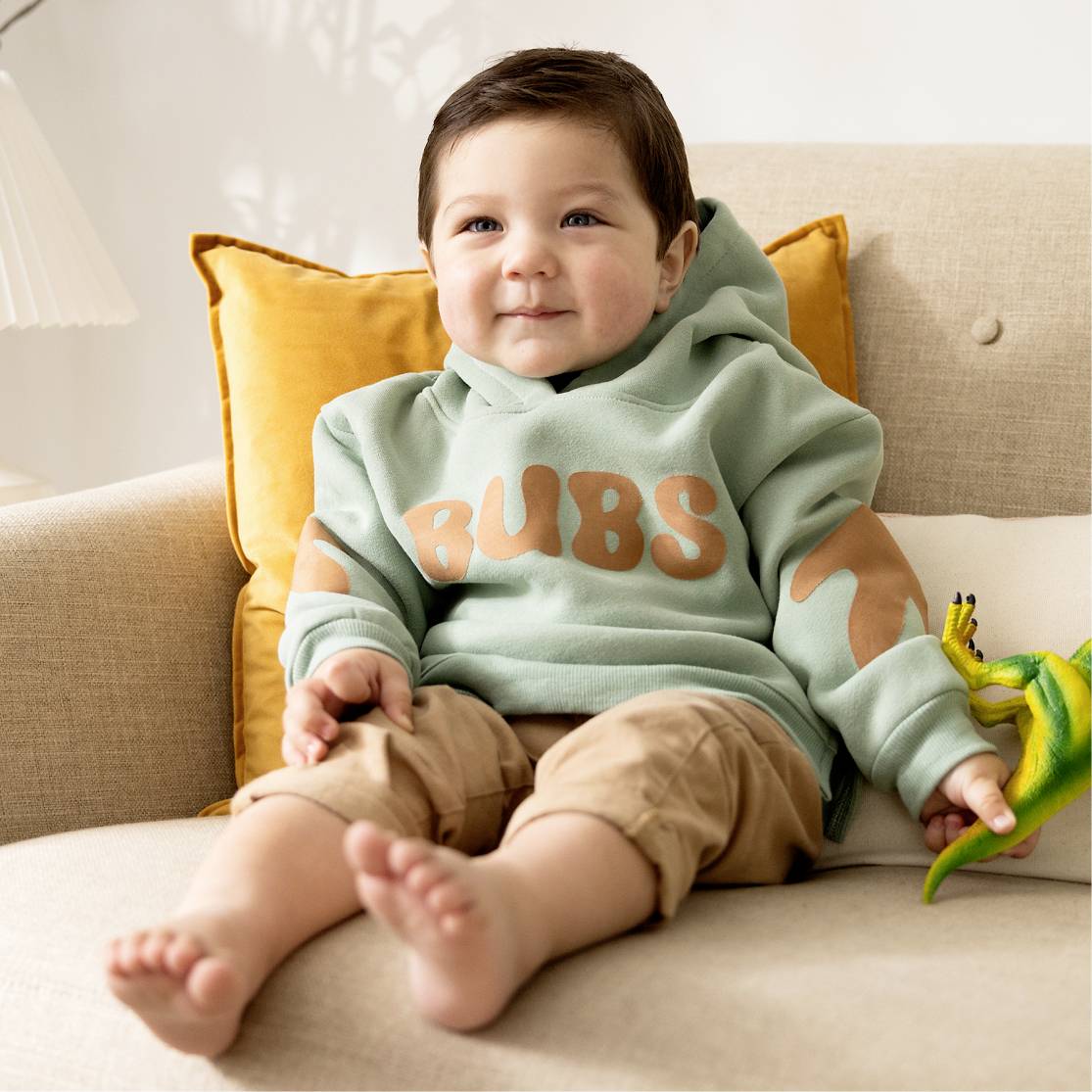 Een blije peuter zit op een bank, draagt een groene hoodie met 'BUBS'-opdruk. Naast hem ligt een speelgoed.