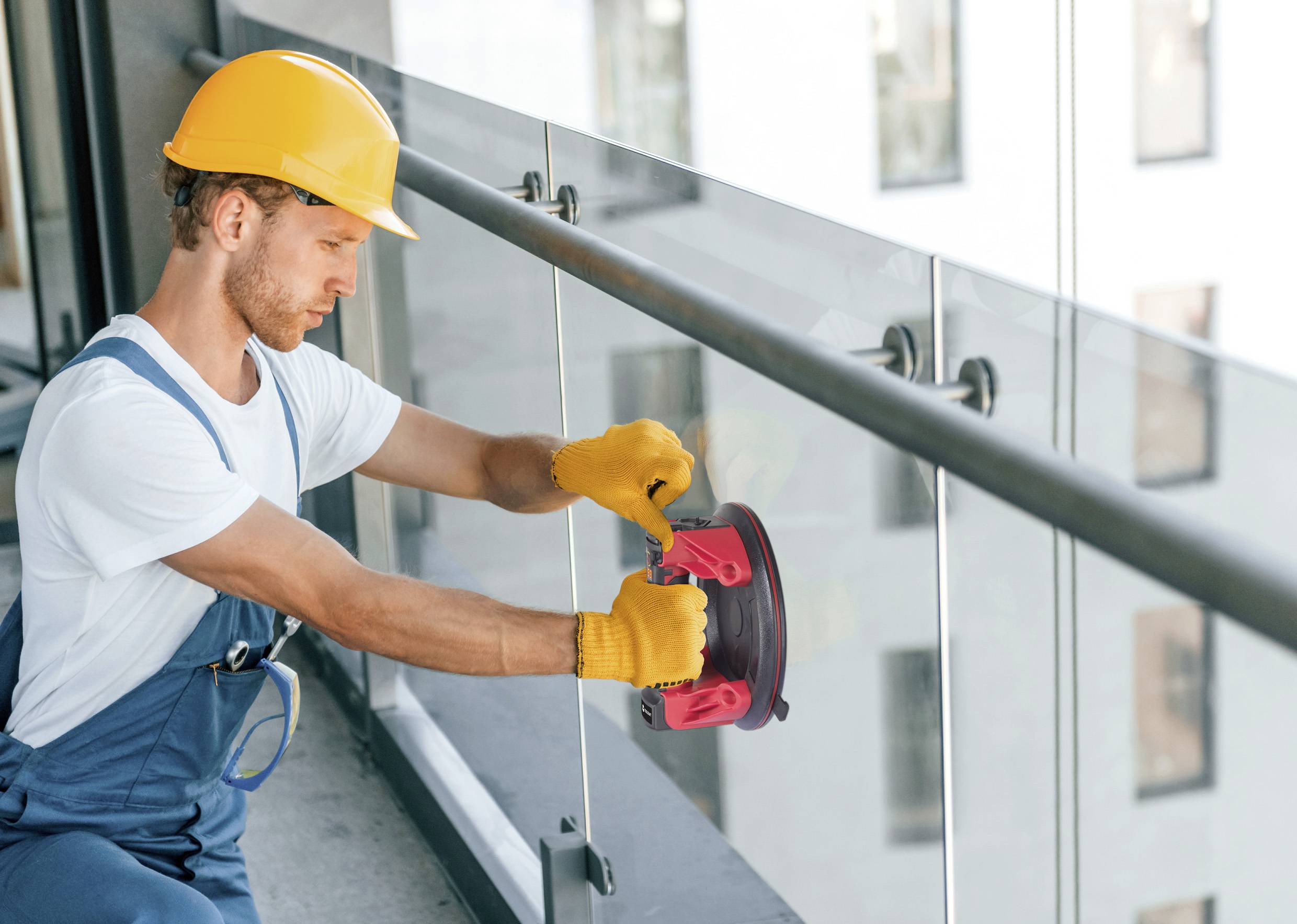 Een arbeider met een gele veiligheidshelm monteert een glazen balustrade op een balkon. Hij draagt handschoenen en gebruikt een gereedschapshouder.