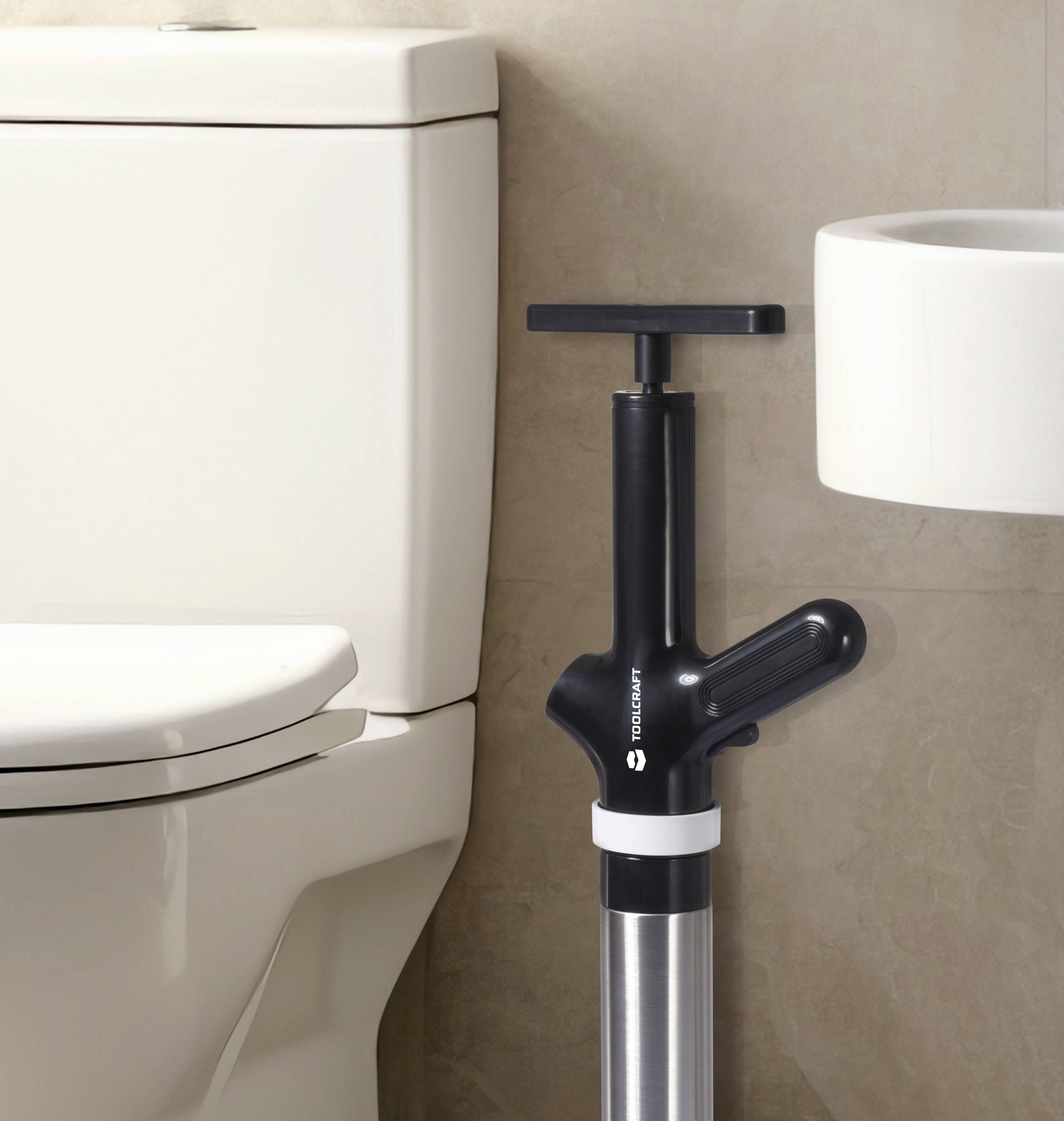 Een toiletpomp staat naast een toilet, klaar voor gebruik. Op de achtergrond een wastafel.