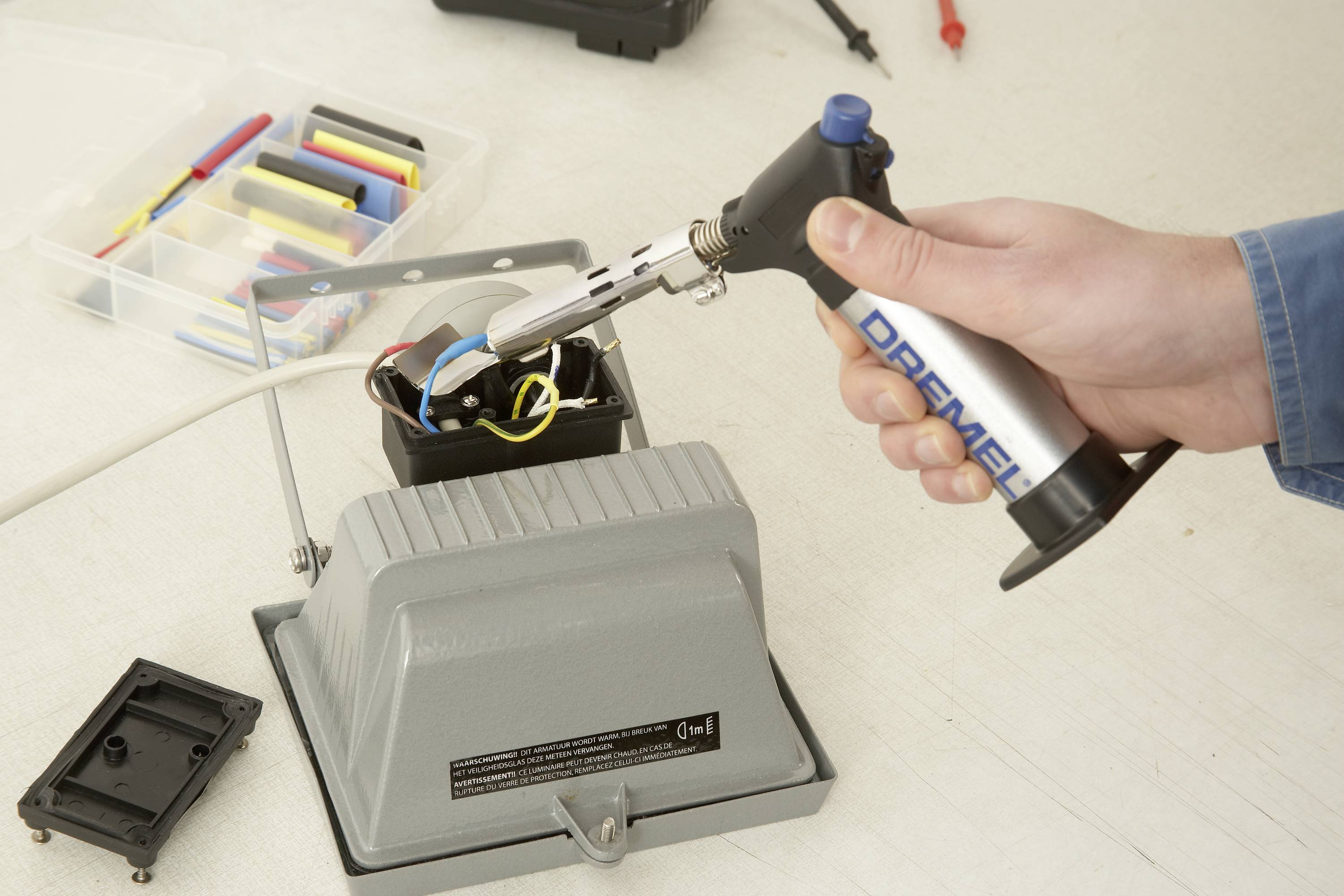 Een persoon gebruikt een soldeerbout om draden aan een elektrische lamp te verbinden. Op de achtergrond is gereedschap zichtbaar.