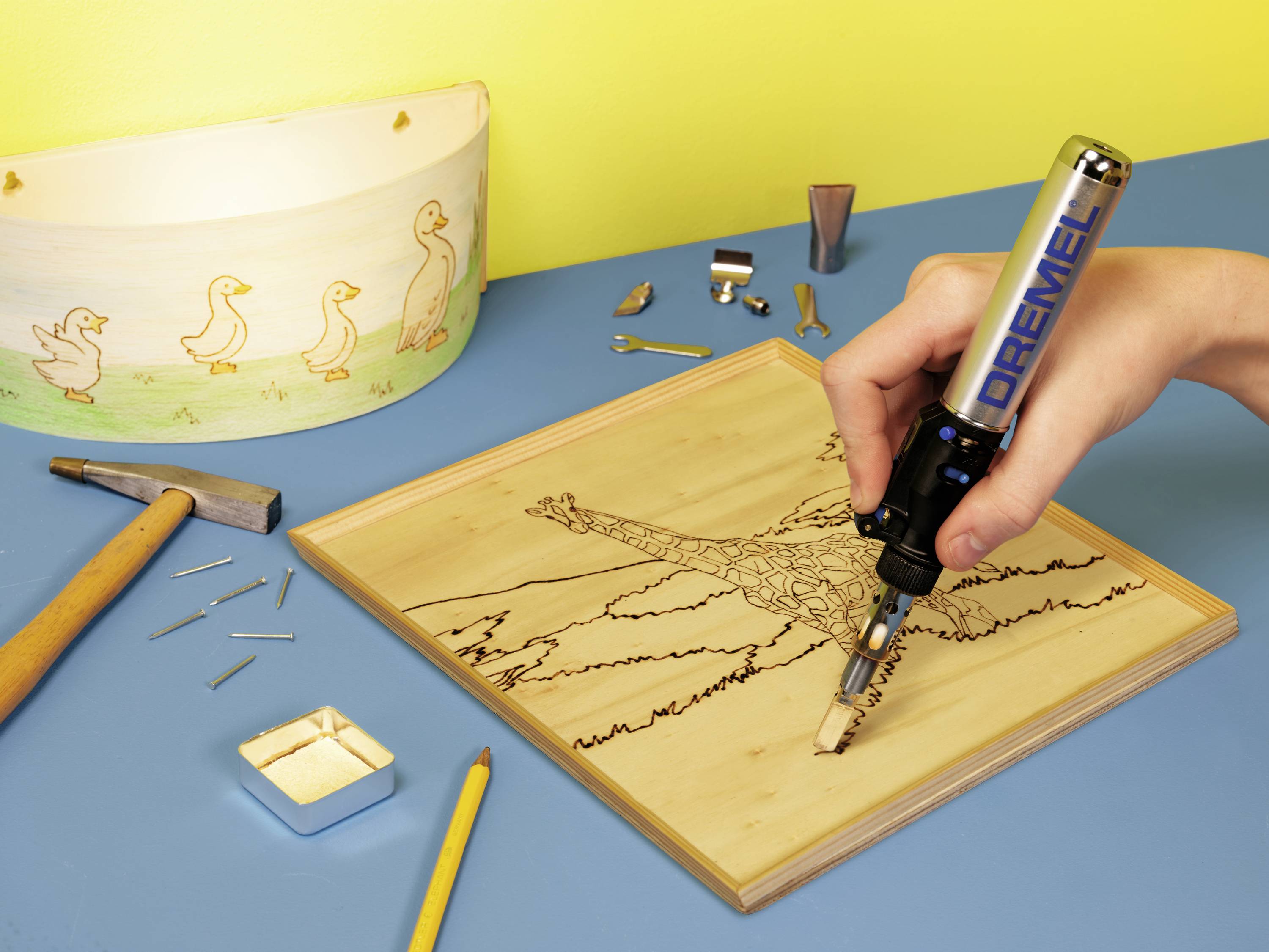 Een hand gebruikt een brandgereedschap om een giraffe in hout te graveren. Op de achtergrond zijn gereedschappen en een beschilderde lampenkap zichtbaar.