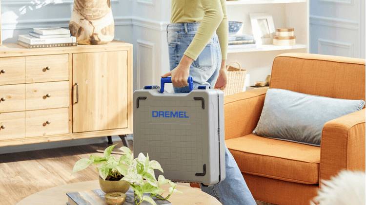 Een persoon die een Dremel gereedschapskoffer draagt in een gezellige, goed verlichte woonkamer met een oranje bank en houten meubels, waarbij een thuisproject-opstelling wordt geaccentueerd.