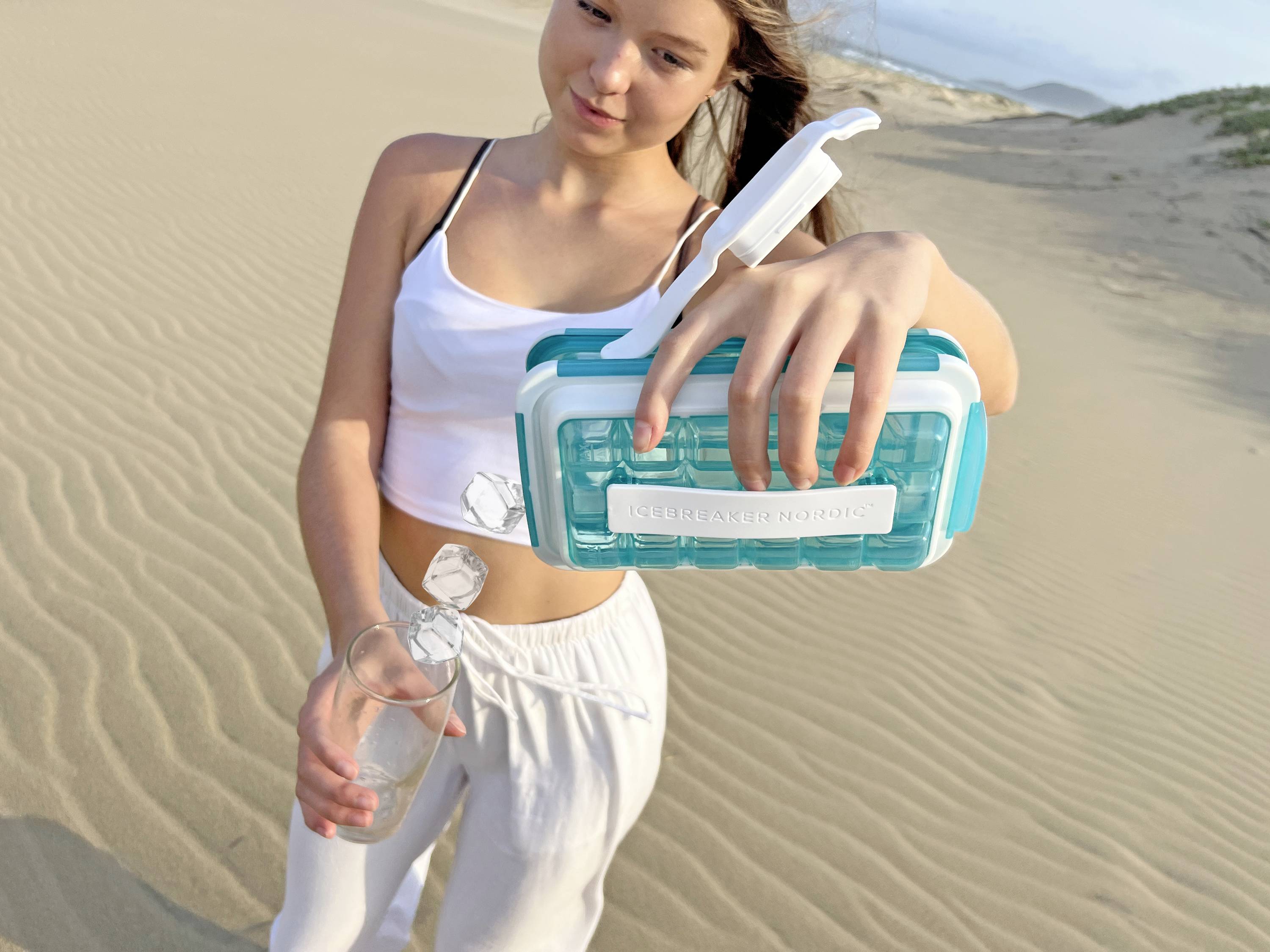 Een vrouw aan een zanderig strand giet water uit een fles in een waterbehoudende container met een geïntegreerd filter.