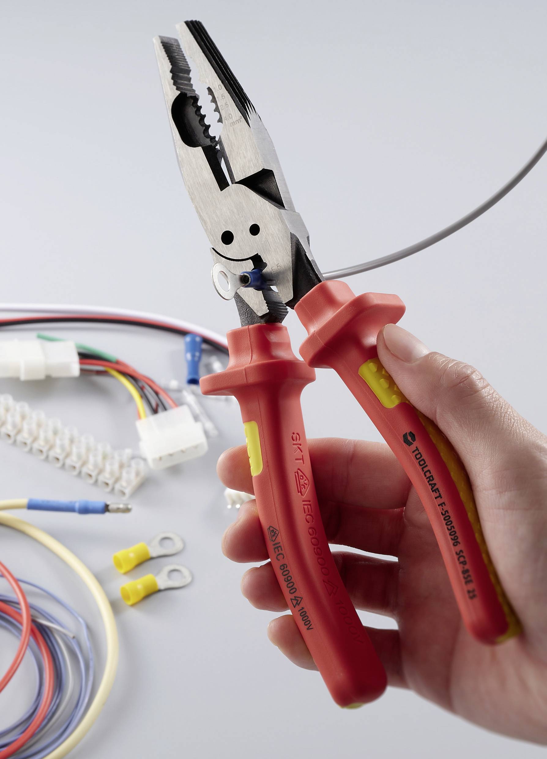 Een hand houdt geïsoleerde combinatietangen met rode handgrepen vast, ter illustratie van gebruik bij elektrisch werk; verschillende kabels en aansluitingen zijn op de achtergrond zichtbaar.