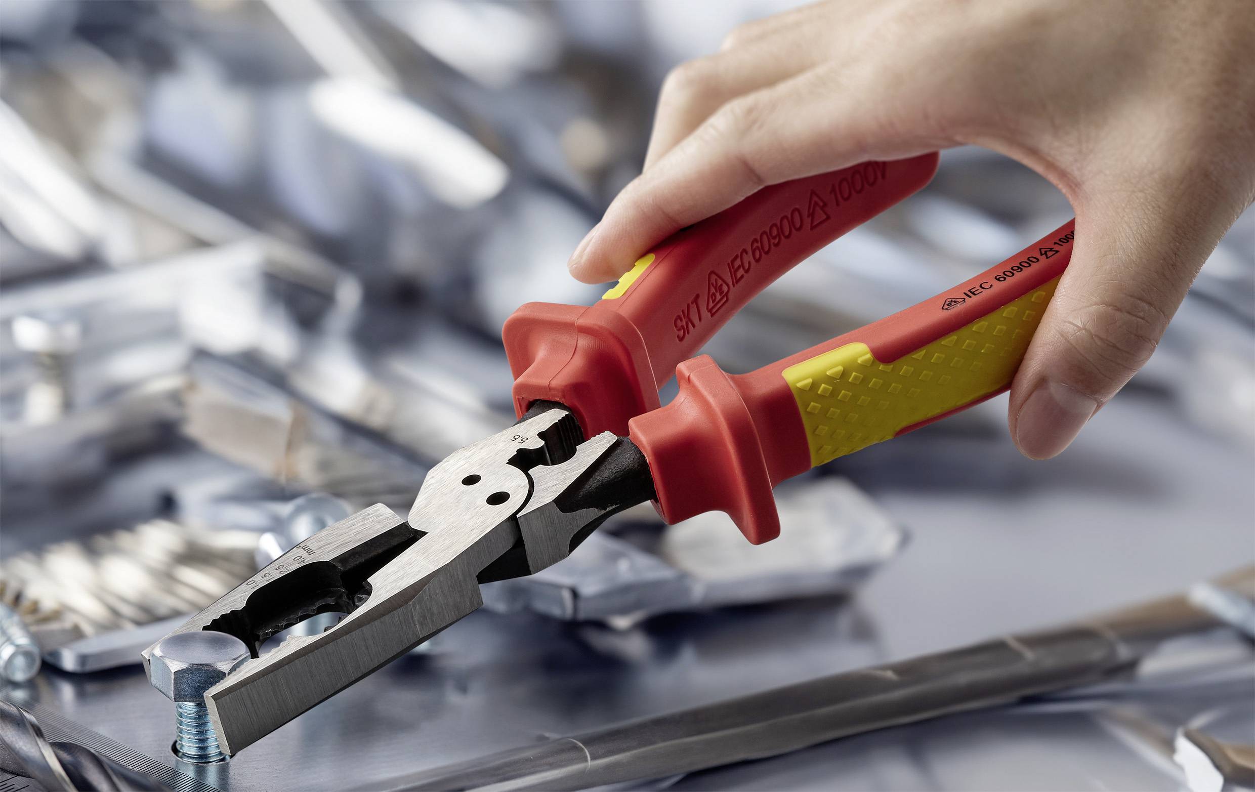 Een hand die rode en gele tangen gebruikt om een bout aan te draaien op een reflecterende metalen oppervlakte, met verspreide metalen onderdelen op de achtergrond.