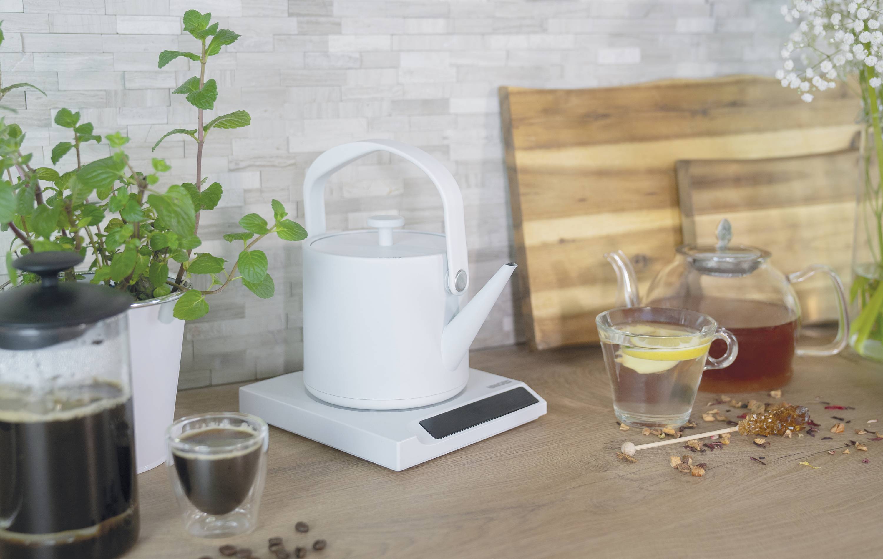 Een witte waterkoker staat op een keukenblad naast een glas thee, een koffiezetter en een pepermuntplant.