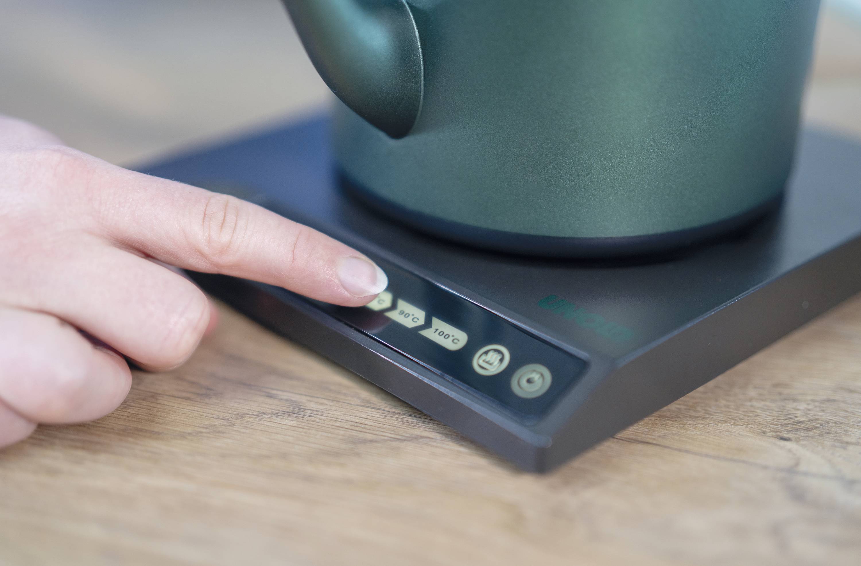 Een hand drukt op een knop van een elektrische kan. De kan staat op een houten oppervlak.