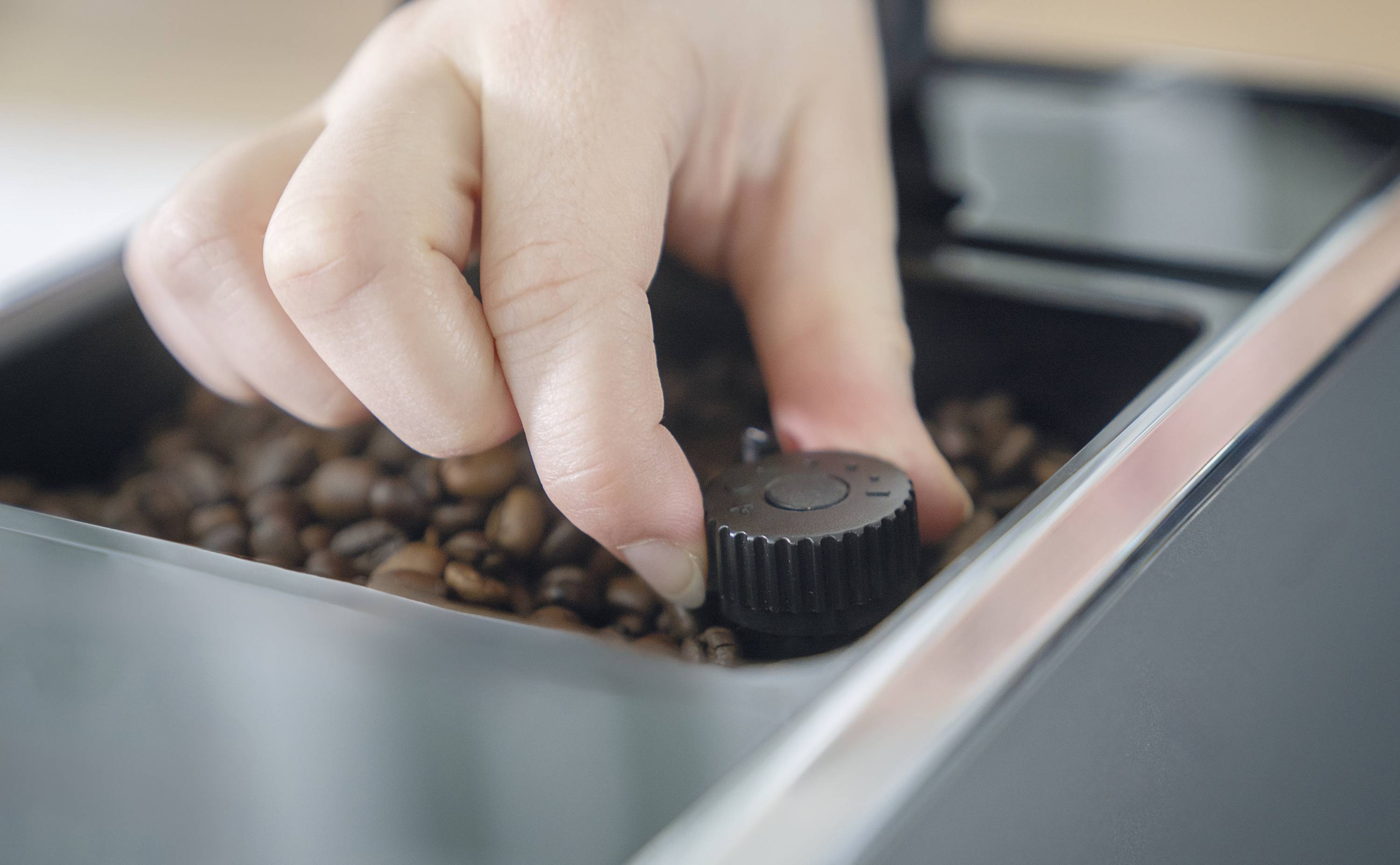 Een hand stelt de maalgraad in bij een koffiemachine met koffiebonen in de houder.