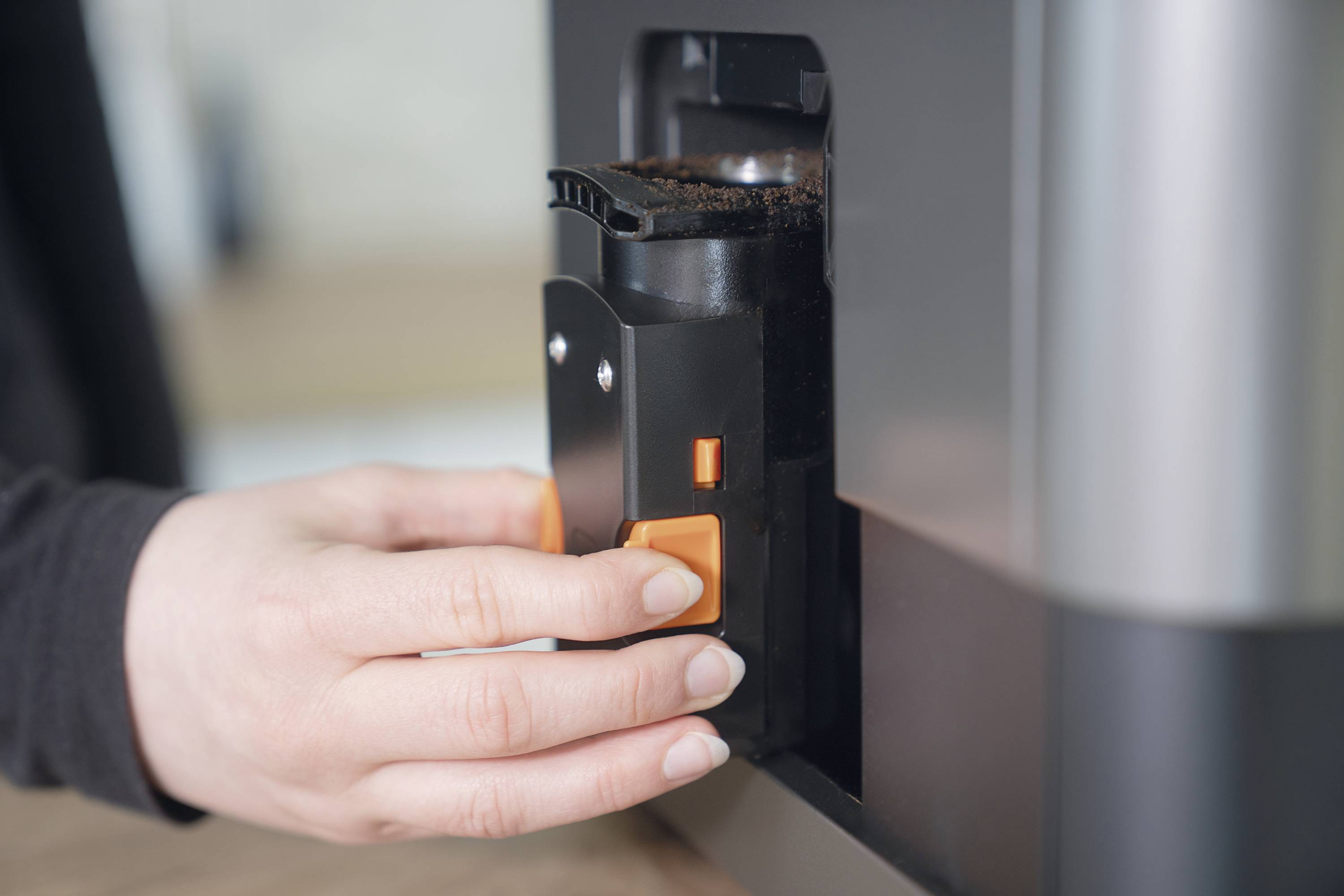 'Persoon houdt een koffiebonenbak bij een koffiemachine, toont het proces van het uitnemen of inzetten van de bak.'