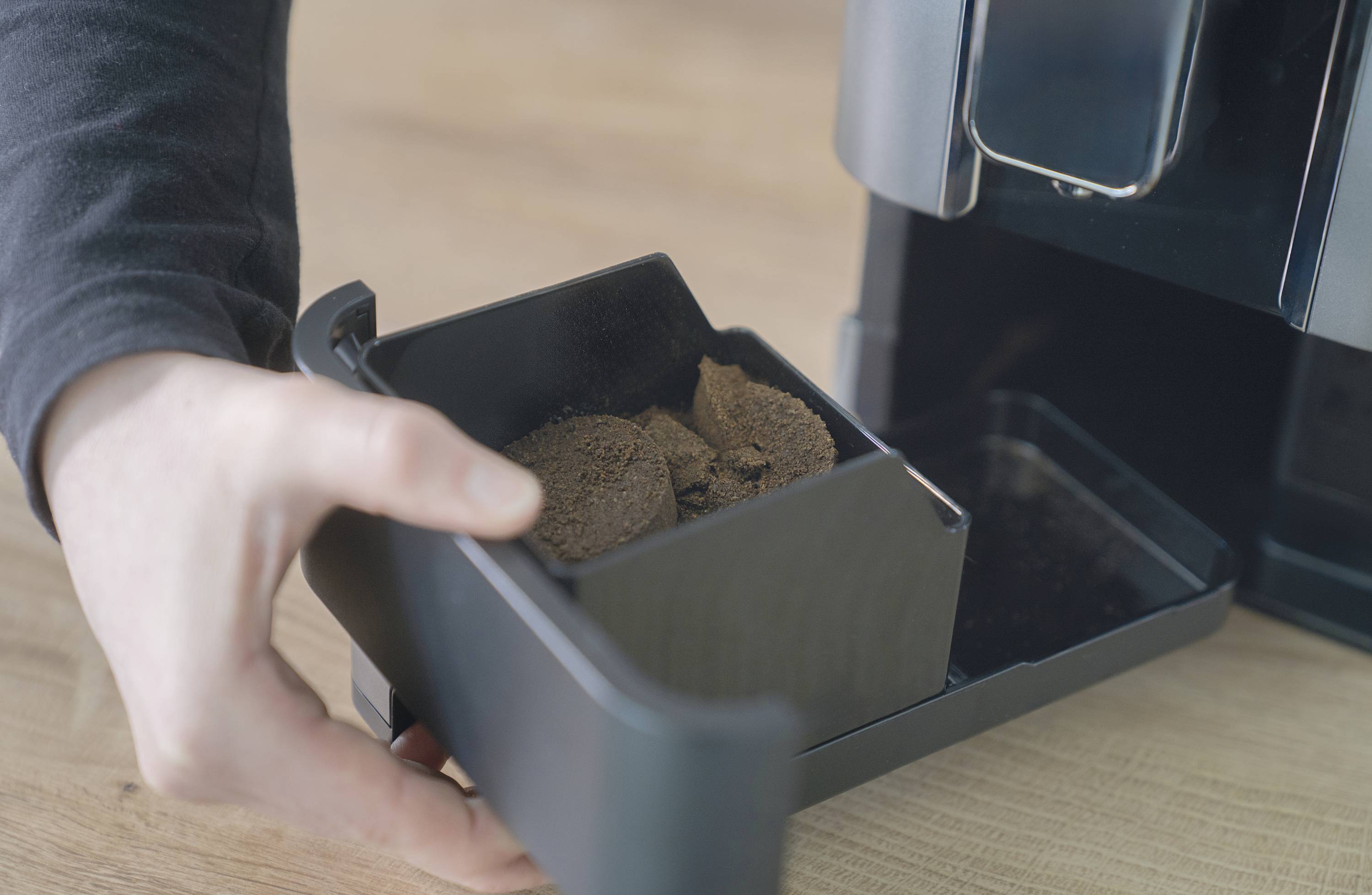 Een hand leegt de koffiedikbak van een koffiemachine op een houten oppervlak.