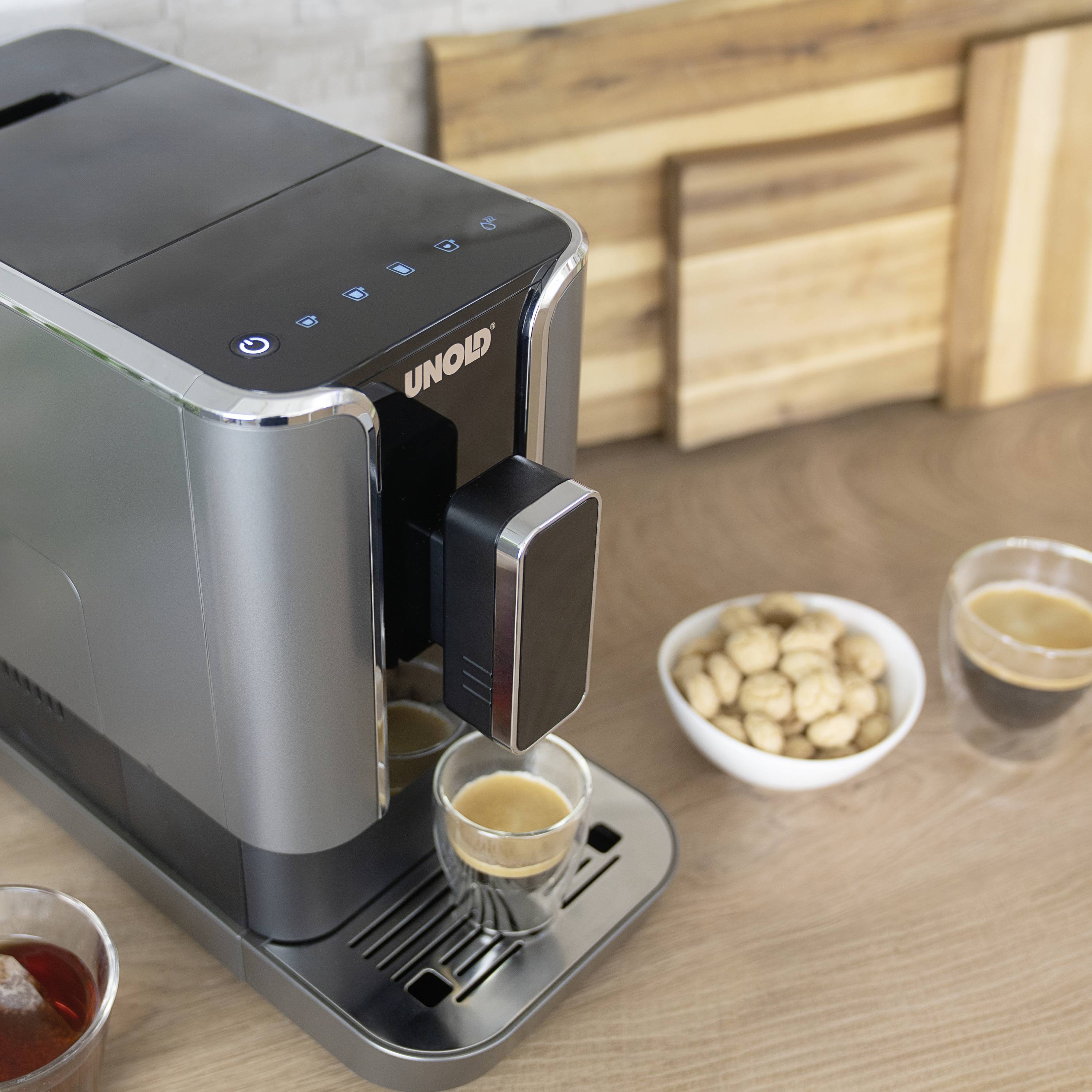 Zwarte koffiemachine op houten werkblad naast twee gevulde espressokopjes en een schaal met koekjes. Houten achtergrond.