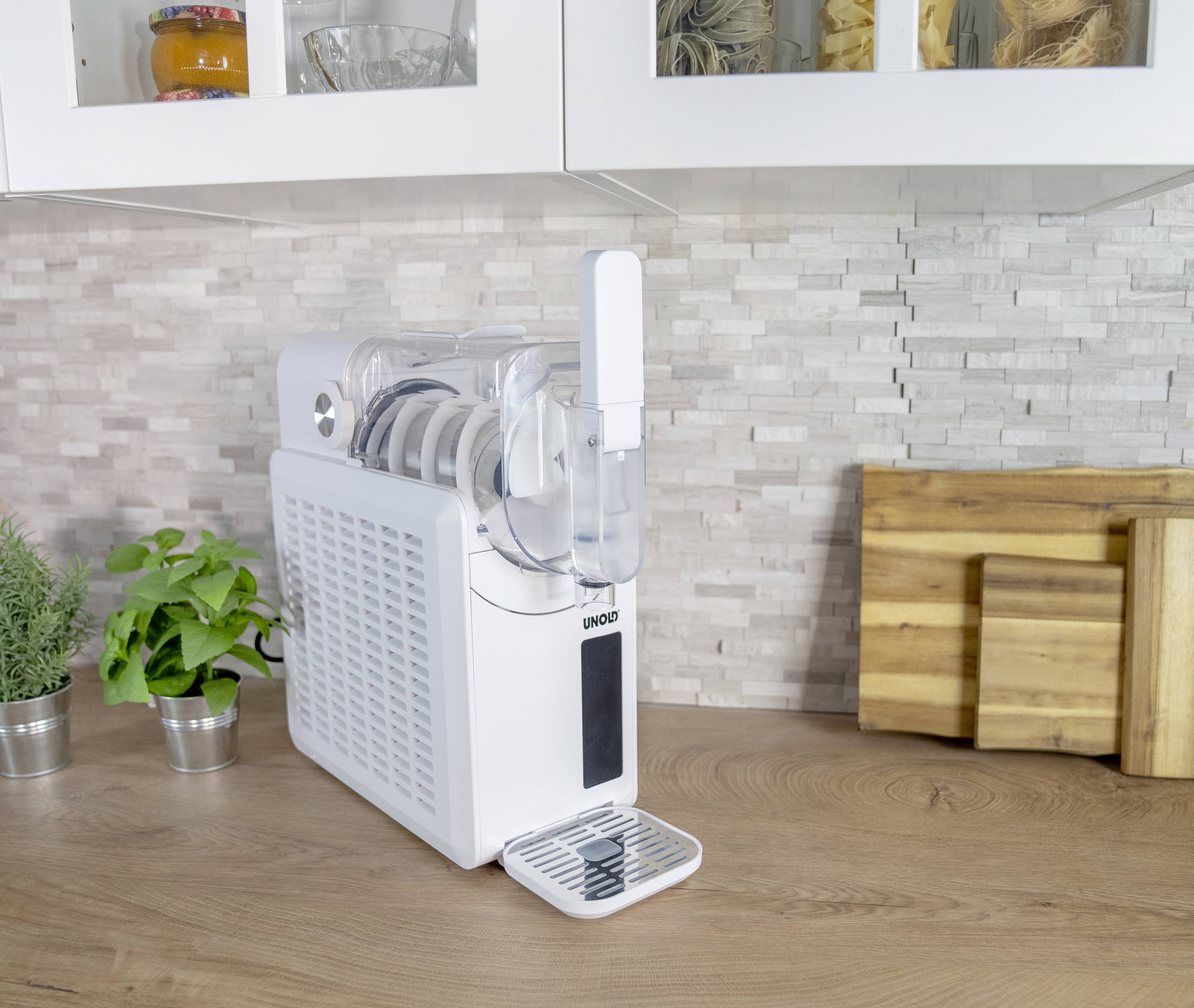 Een witte waterfilter op een aanrecht. Op de achtergrond zijn houten snijplanken, planten en keukengerei zichtbaar.