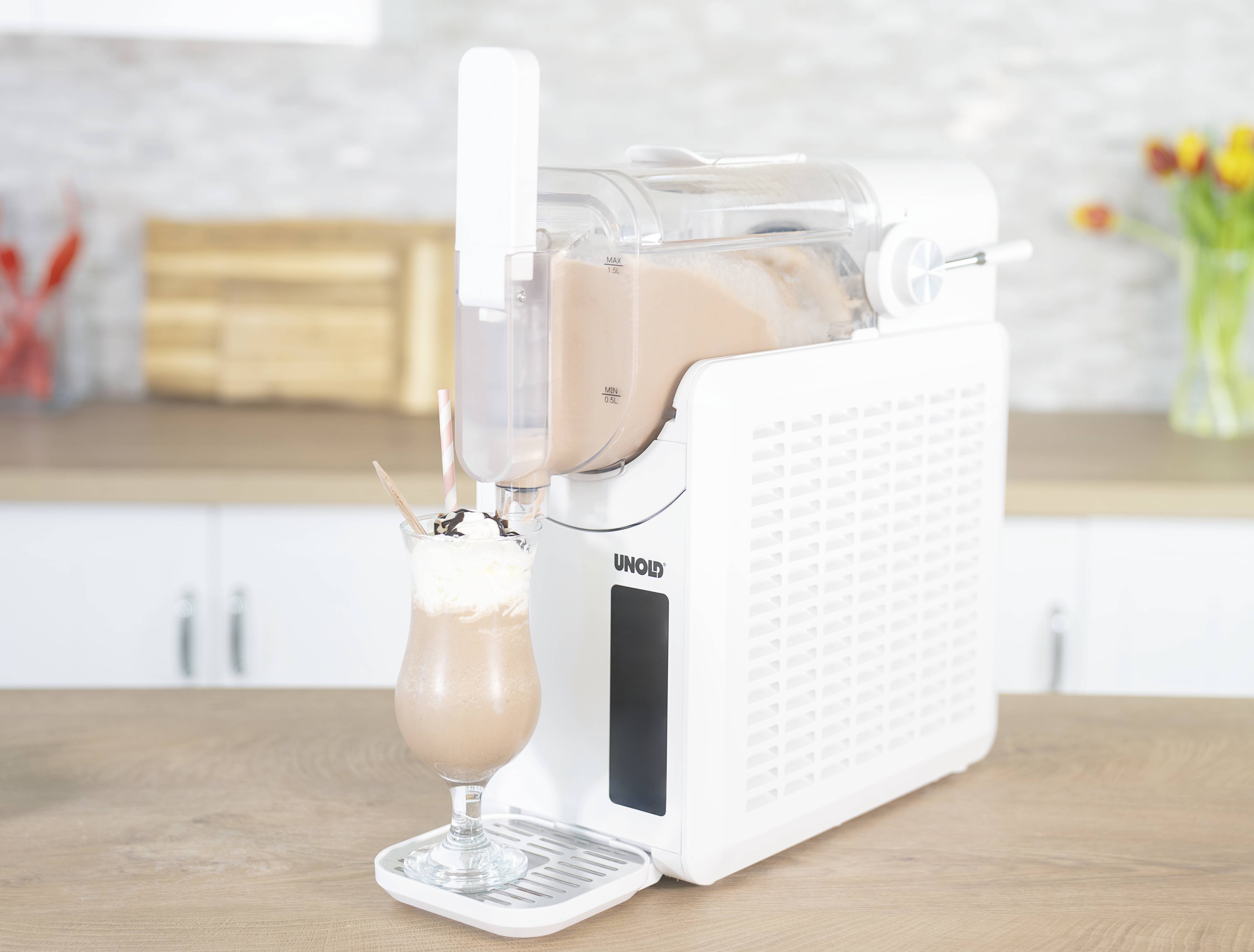 Een ijsmachine op een keukenblad met een klaar ijsdrankje in een glas, daarnaast een boeket gele en rode tulpen.
