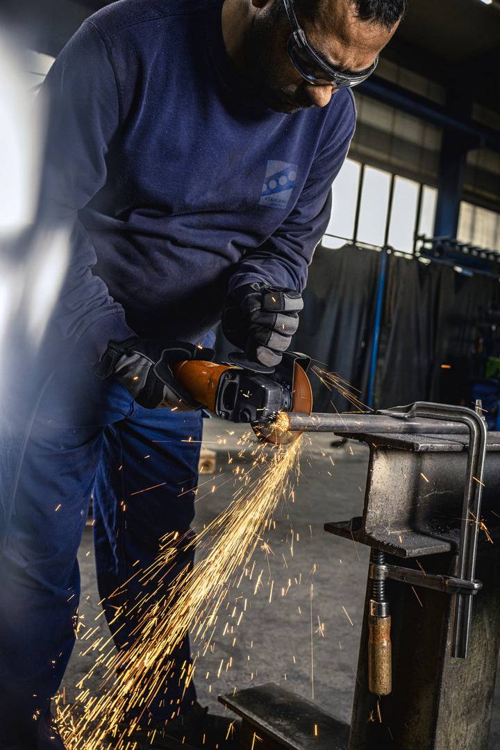 Een arbeider in beschermende uitrusting slijpt een metalen voorwerp met een haakse slijper. Vonken spatten rond, wat wijst op metaalbewerking.