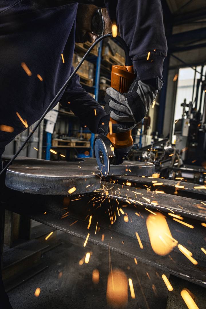 Een ambachtsman slijpt een metalen stuk met een haakse slijpmachine, vonken spatten. Hij draagt beschermende kleding en handschoenen.