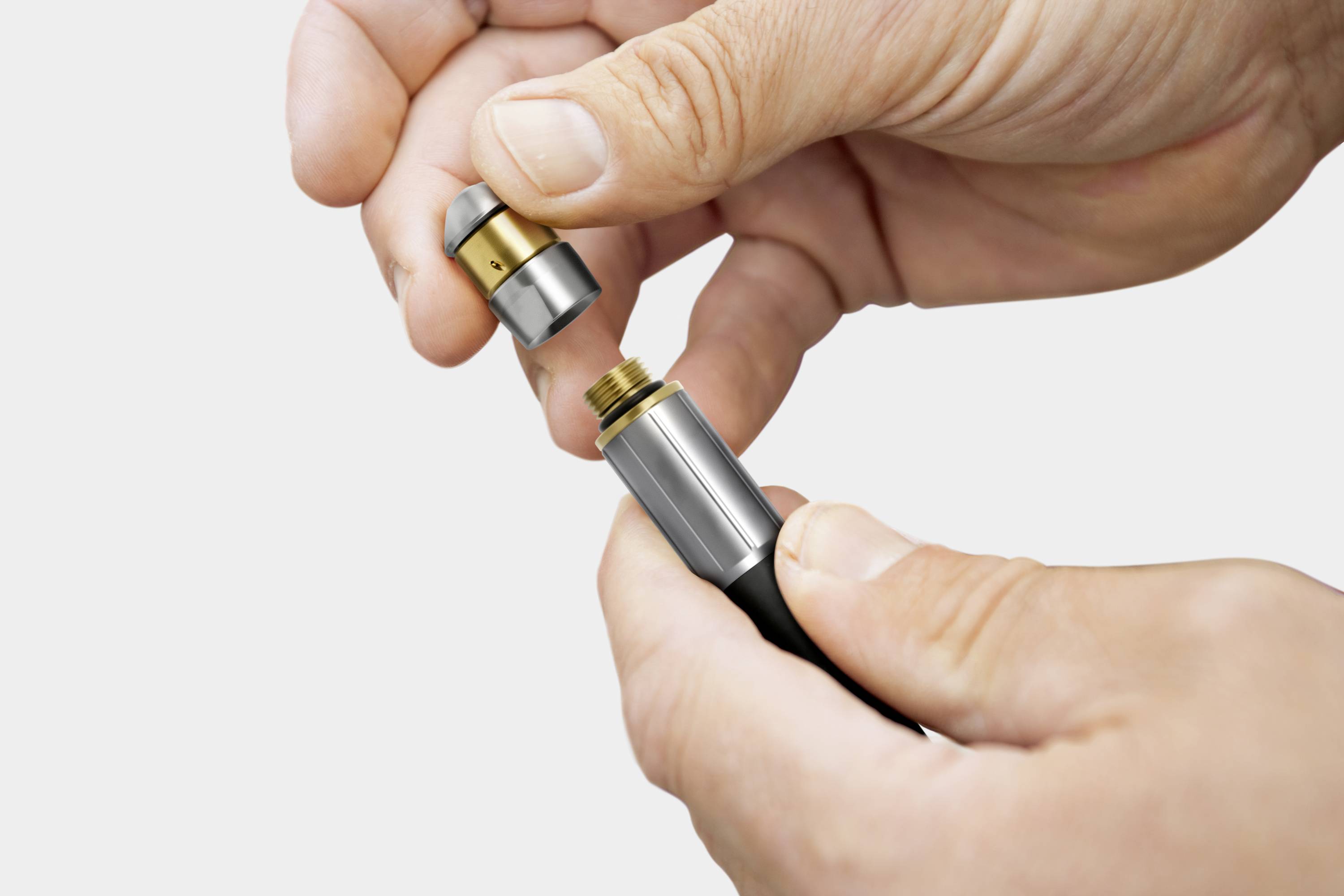 Twee handen monteren een metalen onderdeel op een zwarte voorziening. De afbeelding toont de assemblage van een technisch apparaat.