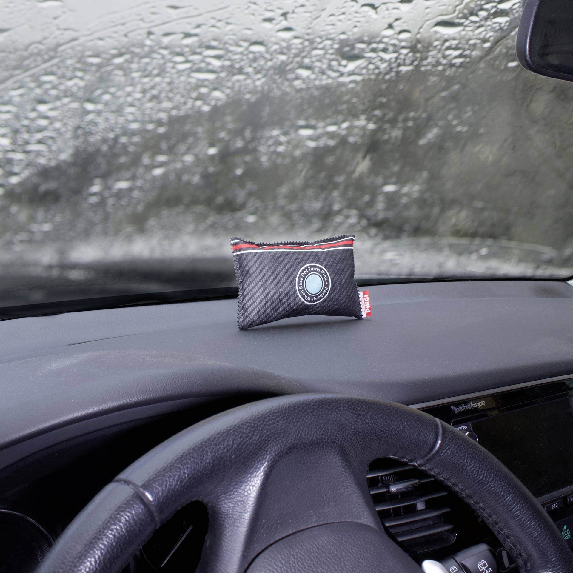 Binnenaanzicht van een auto met een beslagen voorruit; een rechthoekige ontvochtingszak ligt op het dashboard.