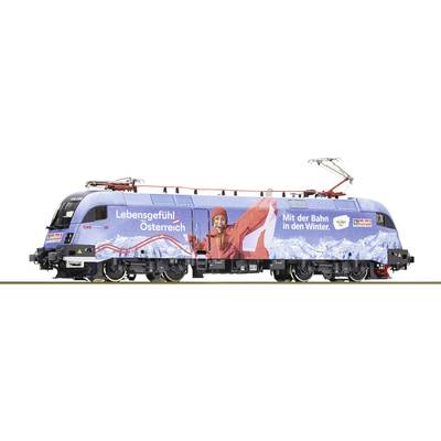 Blauwe locomotief met reclame voor wintertoerisme in Oostenrijk, toont een lachende persoon in rode winterkleding voor bergen.