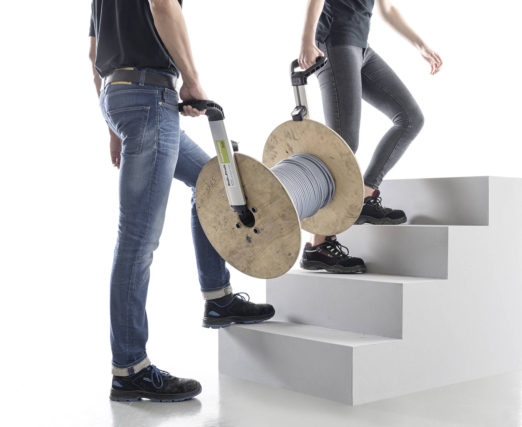 Twee personen dragen een grote kabel- of stroomhaspel met handgrepen een trap op. Ze zijn casual gekleed en de achtergrond is wit.