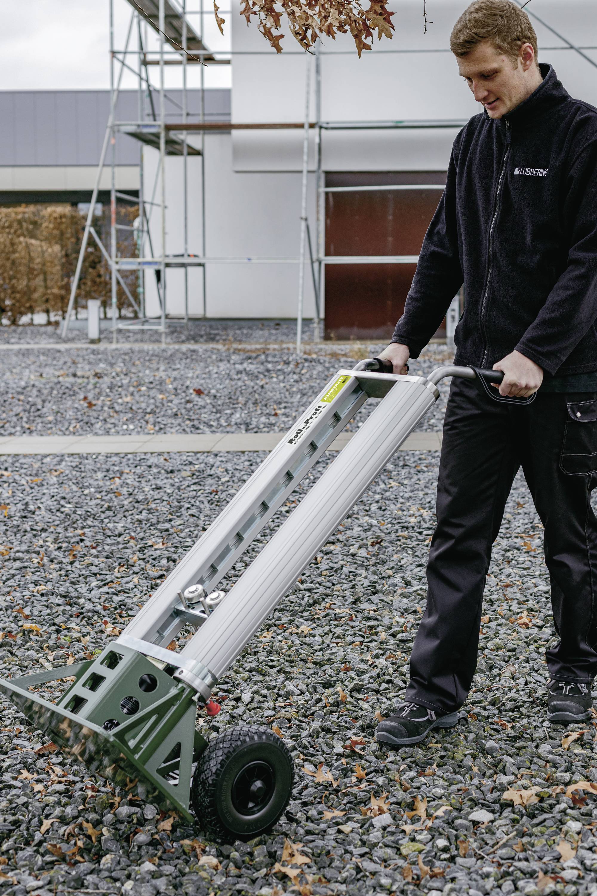 Een man duwt een steekwagen met een lang, rechthoekig voorwerp erop over een grind-ondergrond voor een gebouw.