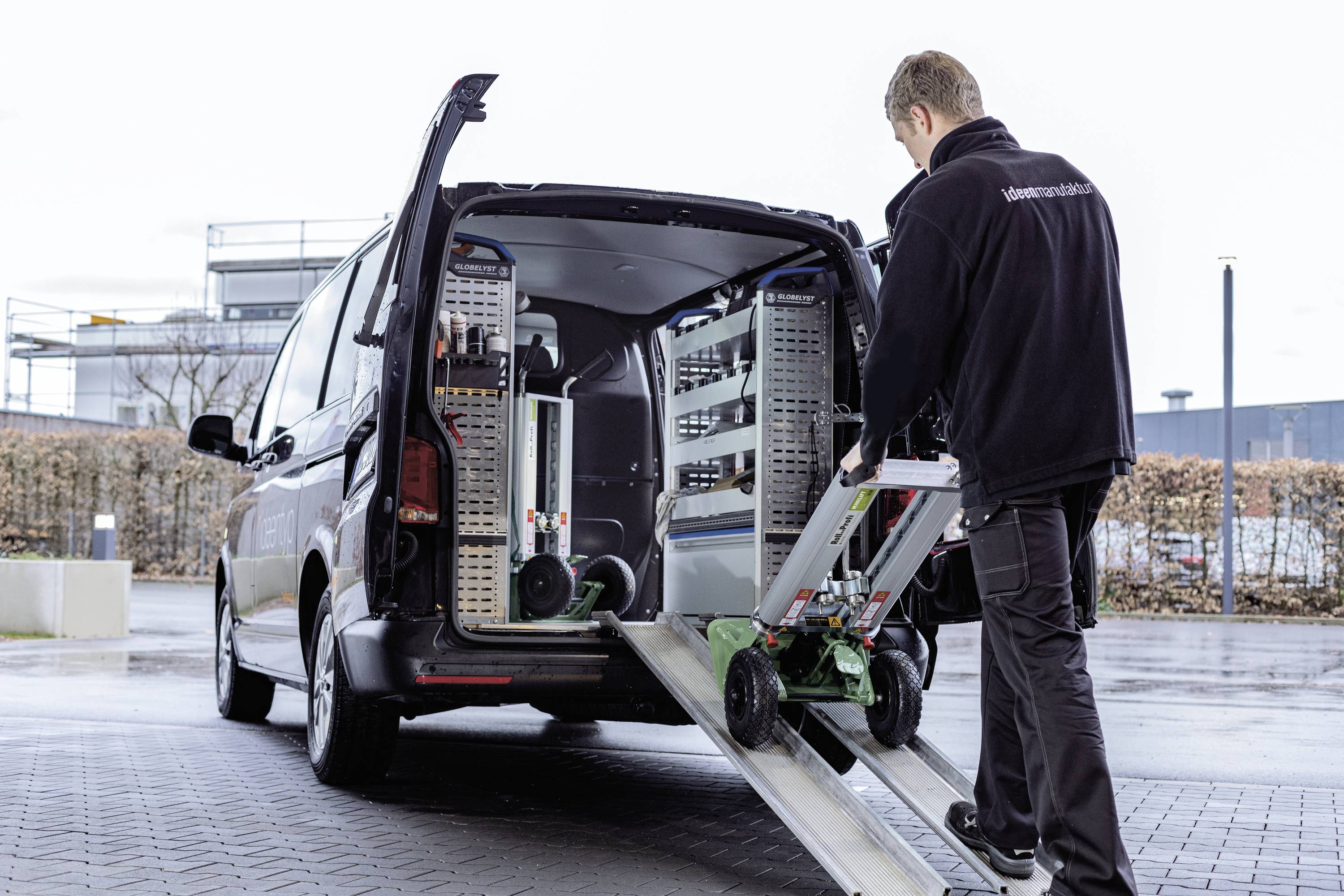Een persoon laadt apparaten op een steekwagen in een bestelbus. Op de achtergrond is een industrieterrein zichtbaar.