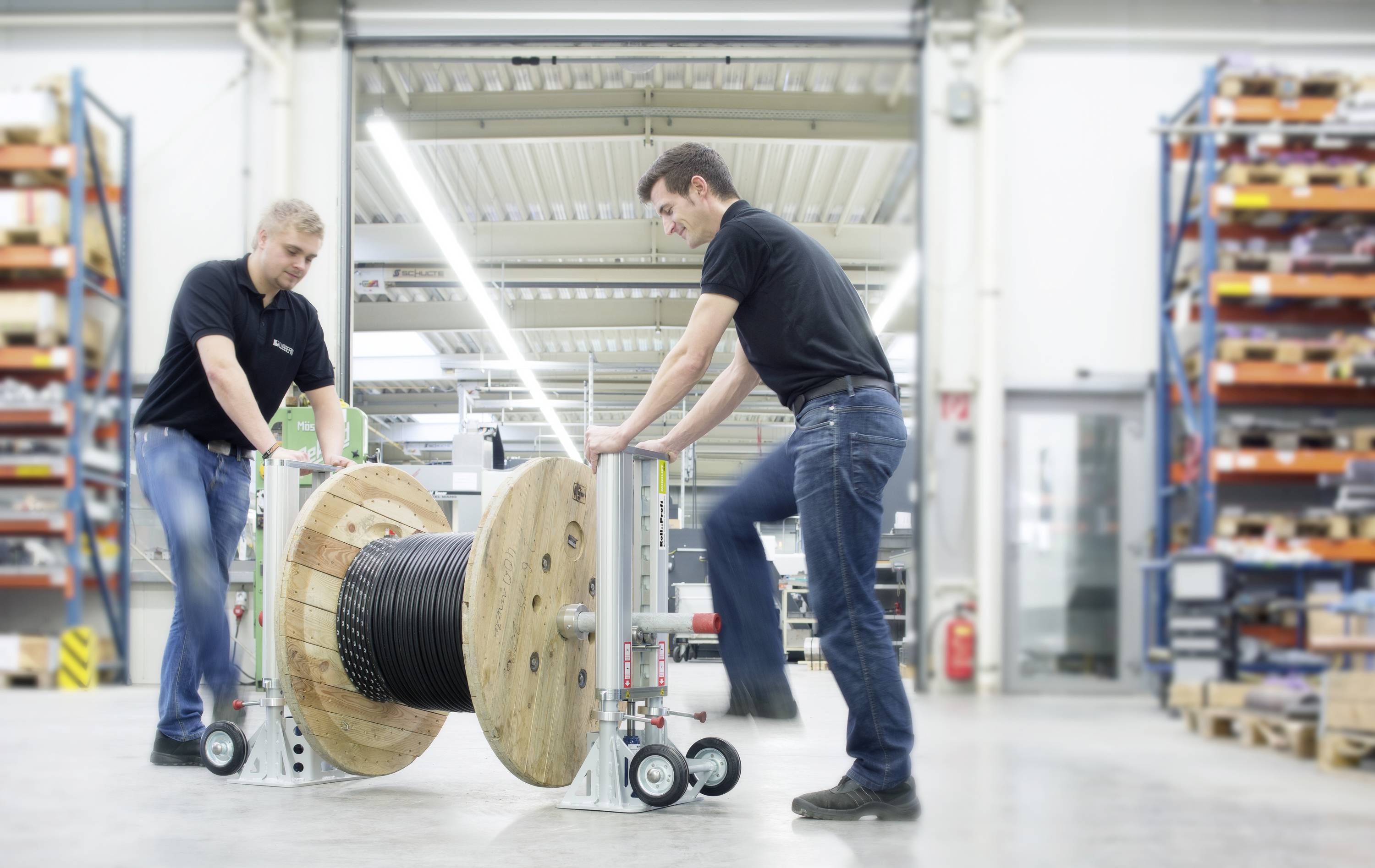 Twee mannen in werkkleding bedienen een grote kabelhaspel in een werkplaats. Ze verplaatsen de haspel met een transportwagen. Op de achtergrond zijn stellingen zichtbaar.